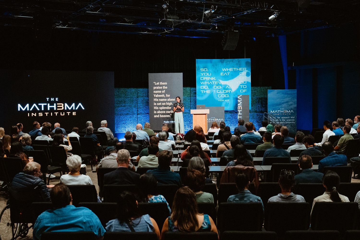 Speaker presents on stage at The Mathema Institute conference with a large audience seated in a darkened auditorium.