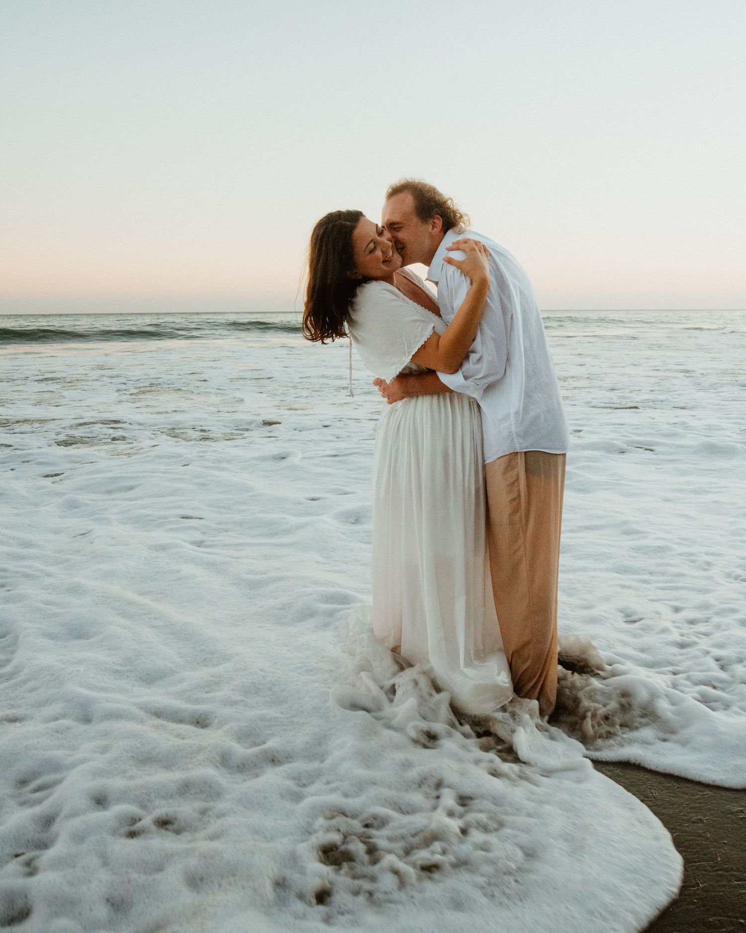 Couple embracing and kissing in white wedding attire as ocean waves wash around them at sunset.