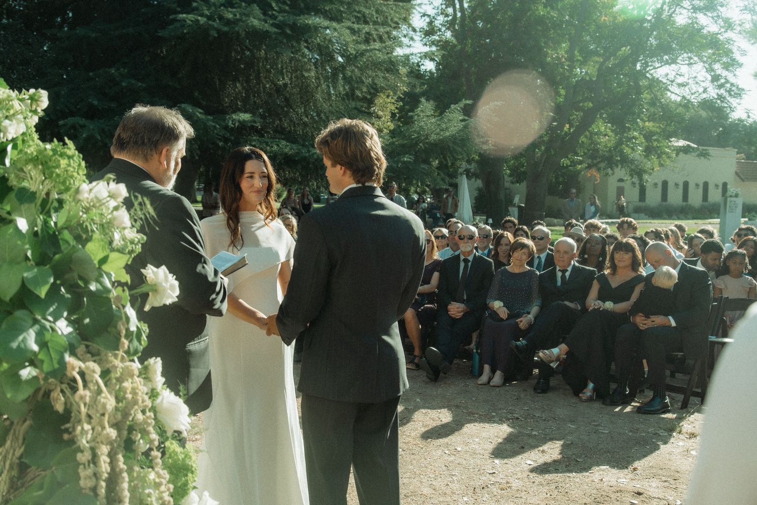 Bride and groom exchanging vows at outdoor wedding ceremony with seated guests in background.