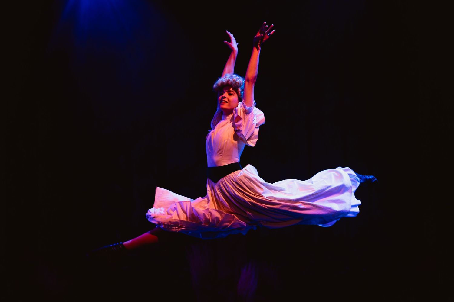 A dancer leaps gracefully in a white flowing costume under blue and purple stage lighting.