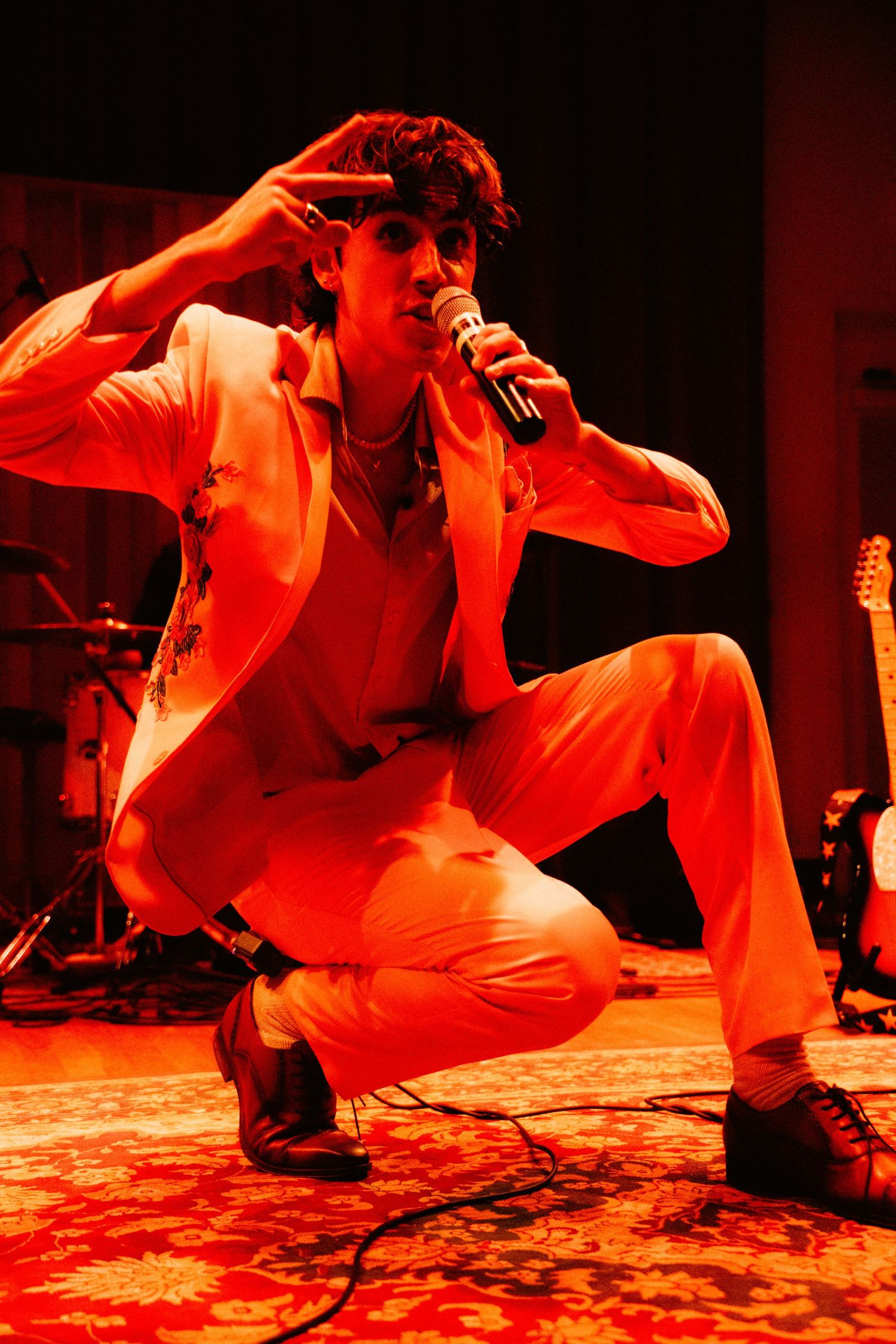 Male performer in white suit crouching on stage under red lights holding microphone during concert.