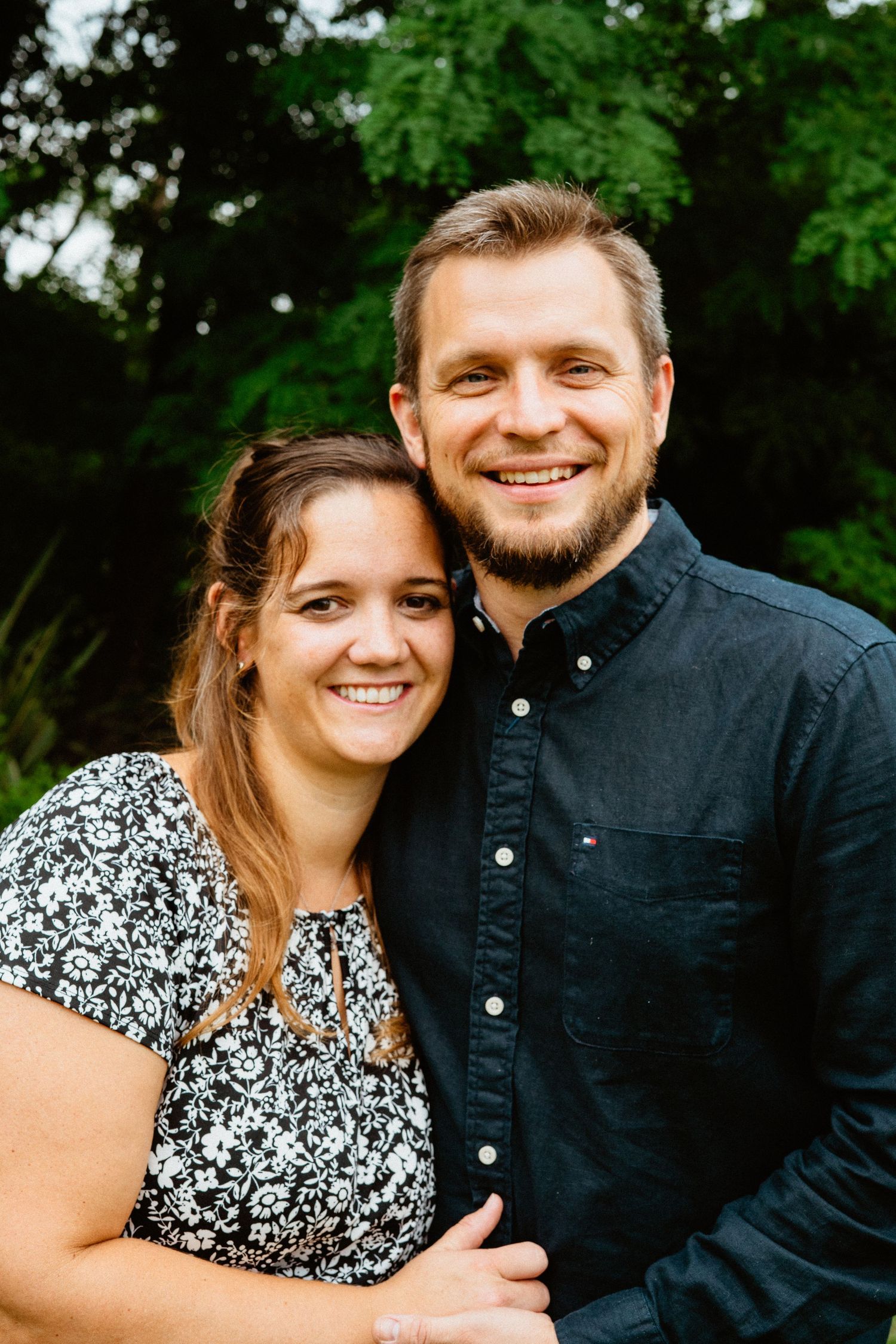 Smiling couple posing together outdoors with green foliage in background.