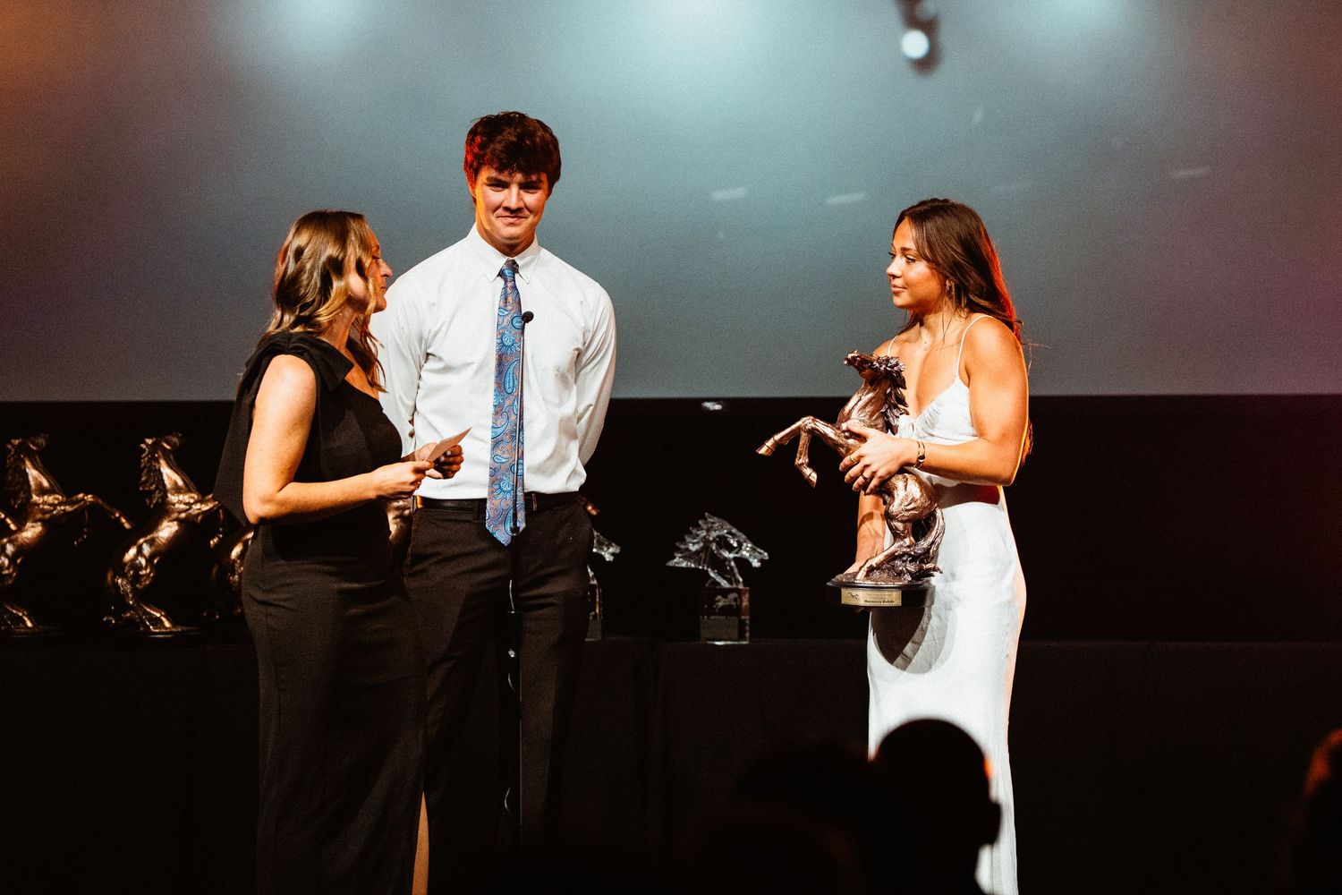 Three people on stage at an awards ceremony, with a woman holding a trophy in a white dress.