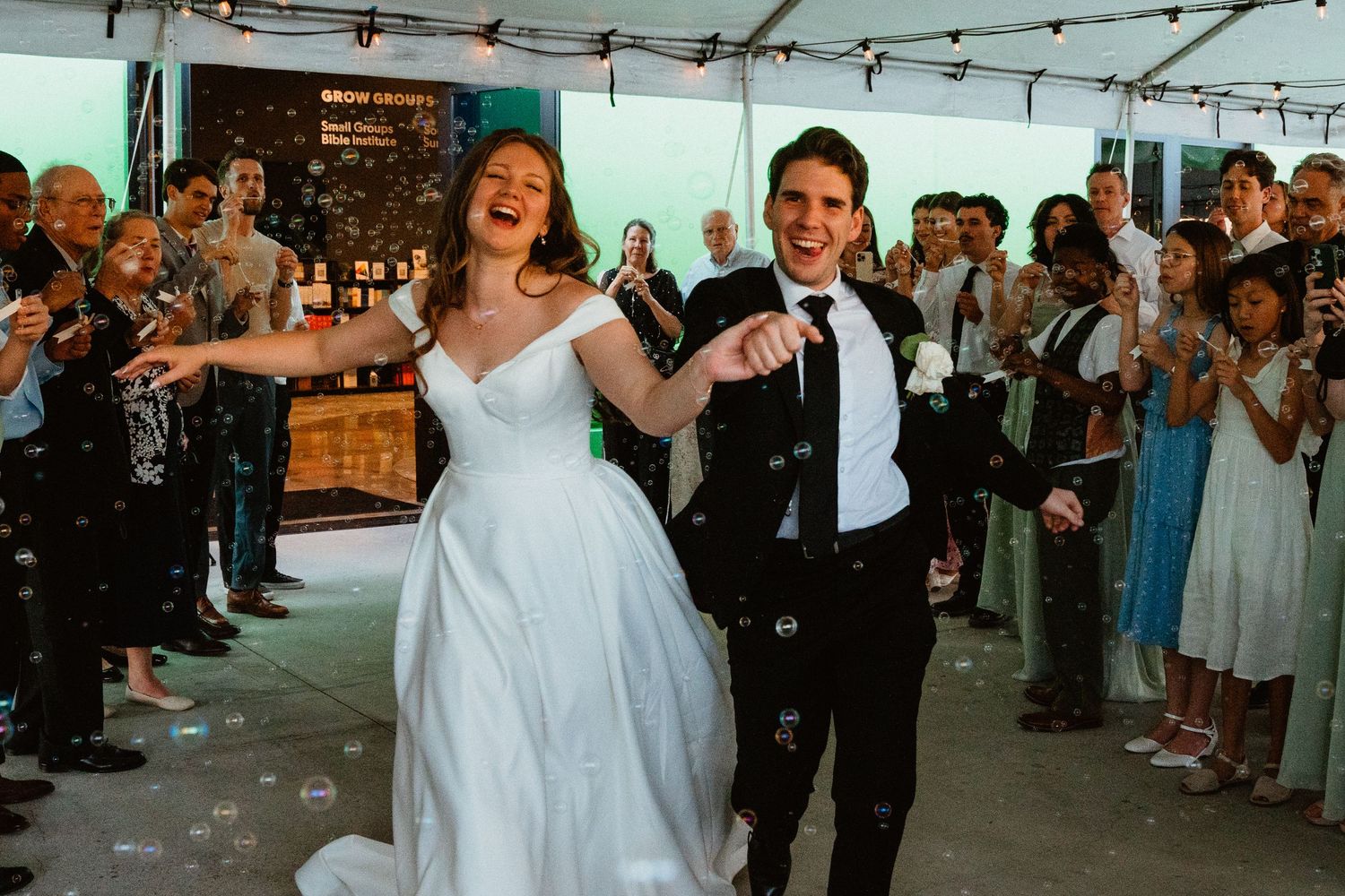 Joyful newlyweds walk hand-in-hand through bubbles while guests celebrate at their outdoor wedding reception.
