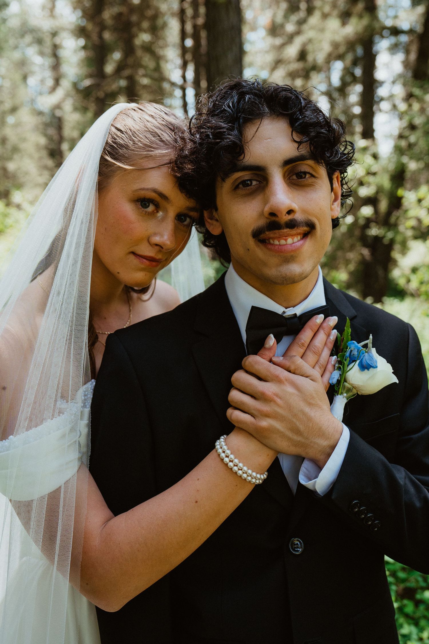 Bride and groom pose intimately in forest, she wears veil and pearl bracelet, he sports a mustache and boutonniere.