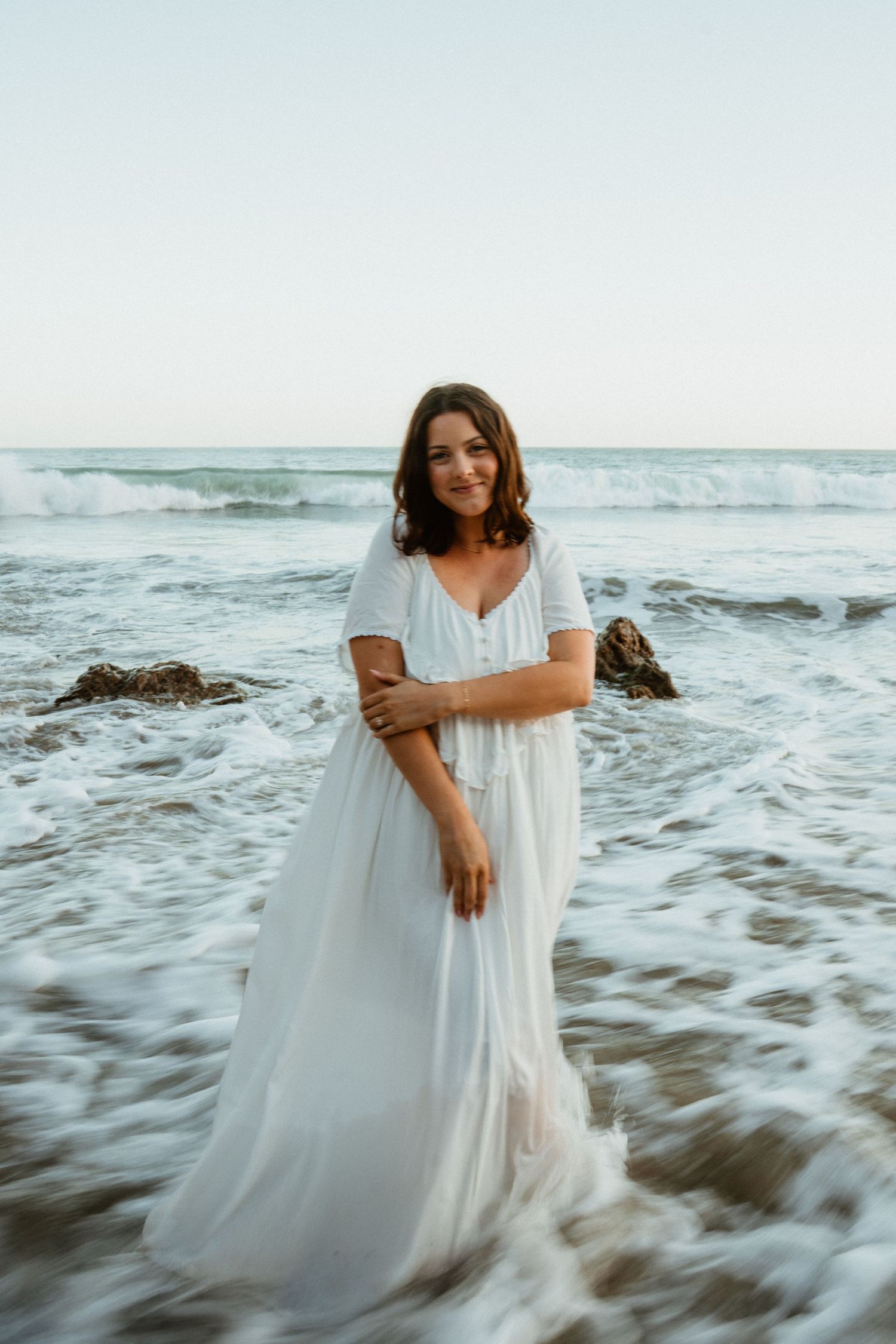 Woman in white dress standing in ocean waves at beach during golden hour.