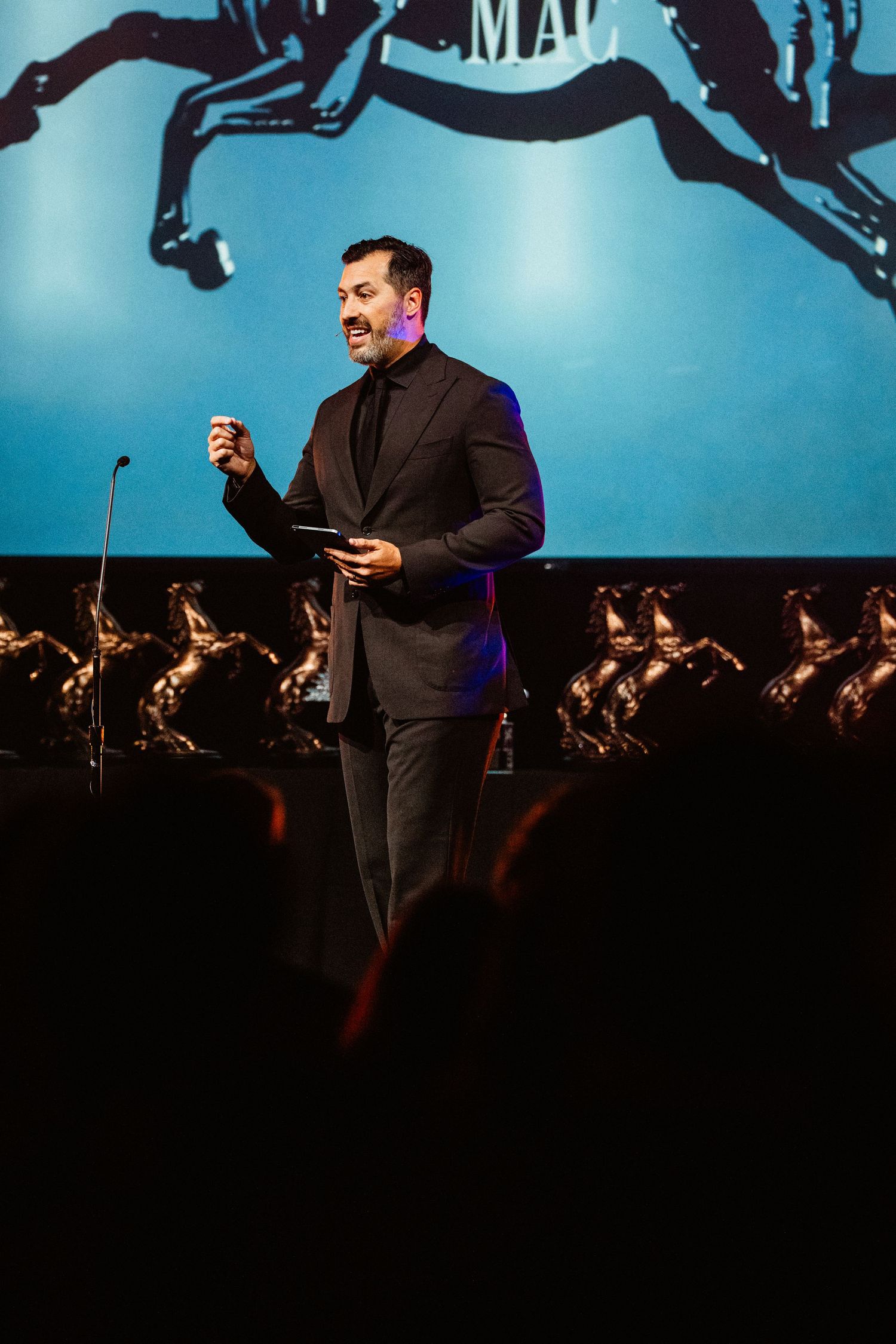 A man in a dark suit speaks on stage holding a microphone with a blue projection screen behind him.