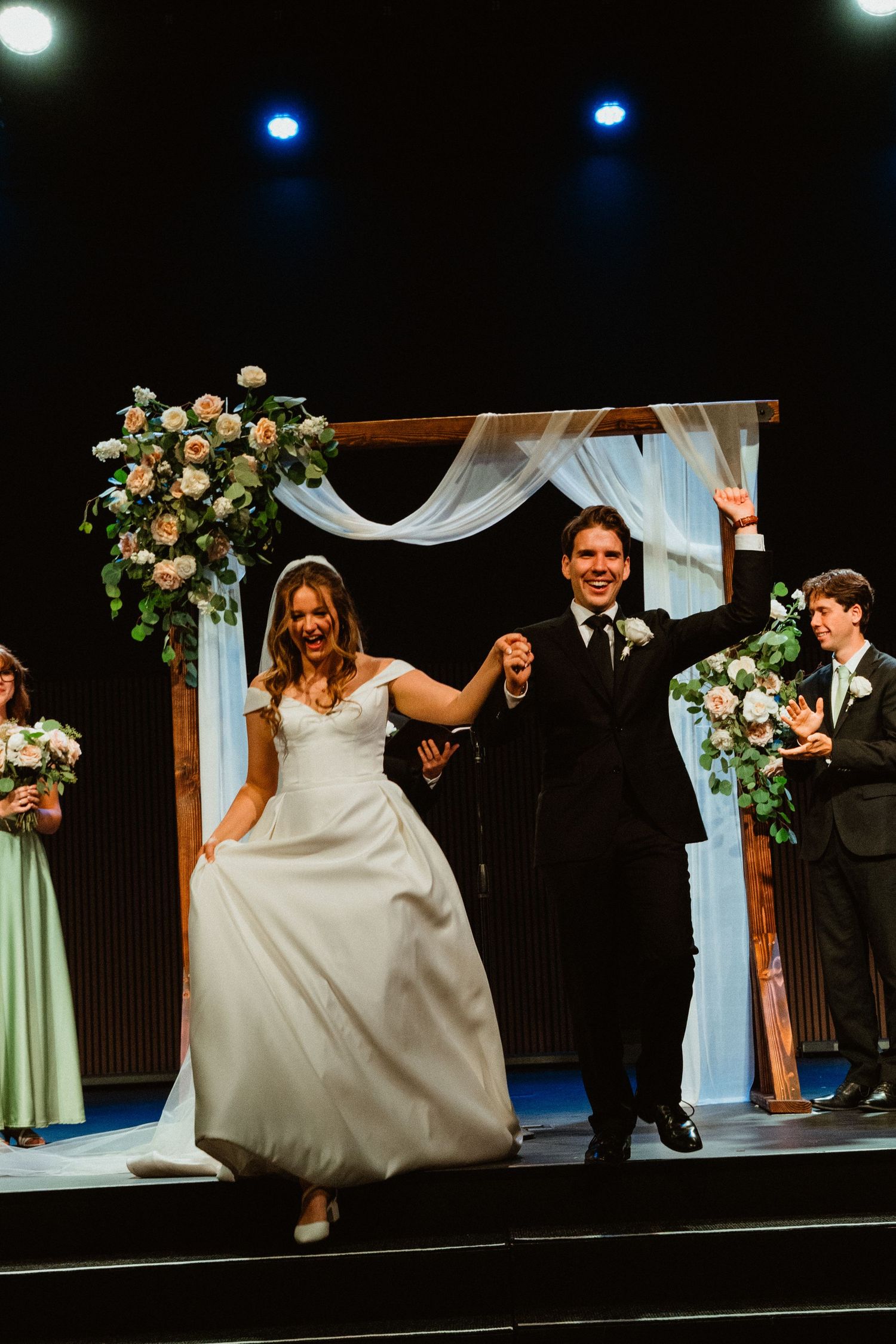 Happy newlyweds celebrating as they walk down the aisle under a floral wooden arch at their wedding.