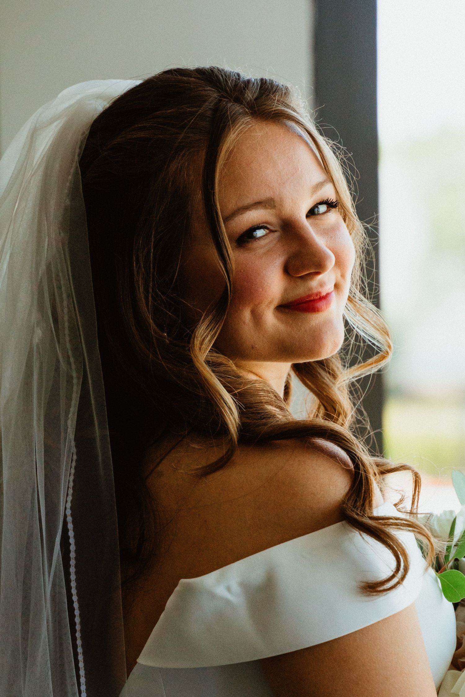 Smiling bride in white off-shoulder gown and veil, holding bouquet, posing elegantly near a window.