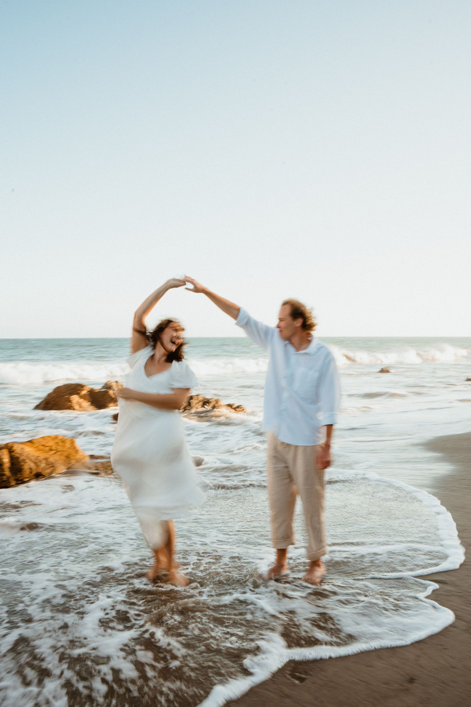 Couple dancing barefoot on a sandy beach at sunset, woman in white dress being twirled by man in white shirt.