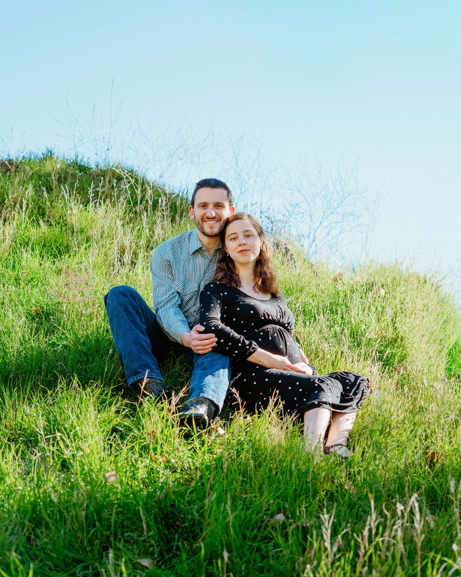 Couple sitting together on a green grassy hillside under clear sky.