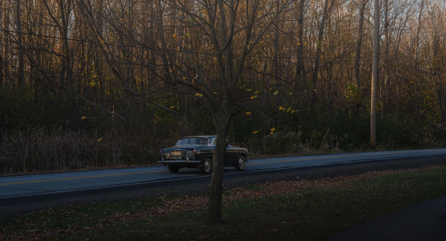 Vintage black truck driving on a quiet road surrounded by autumn forest trees.