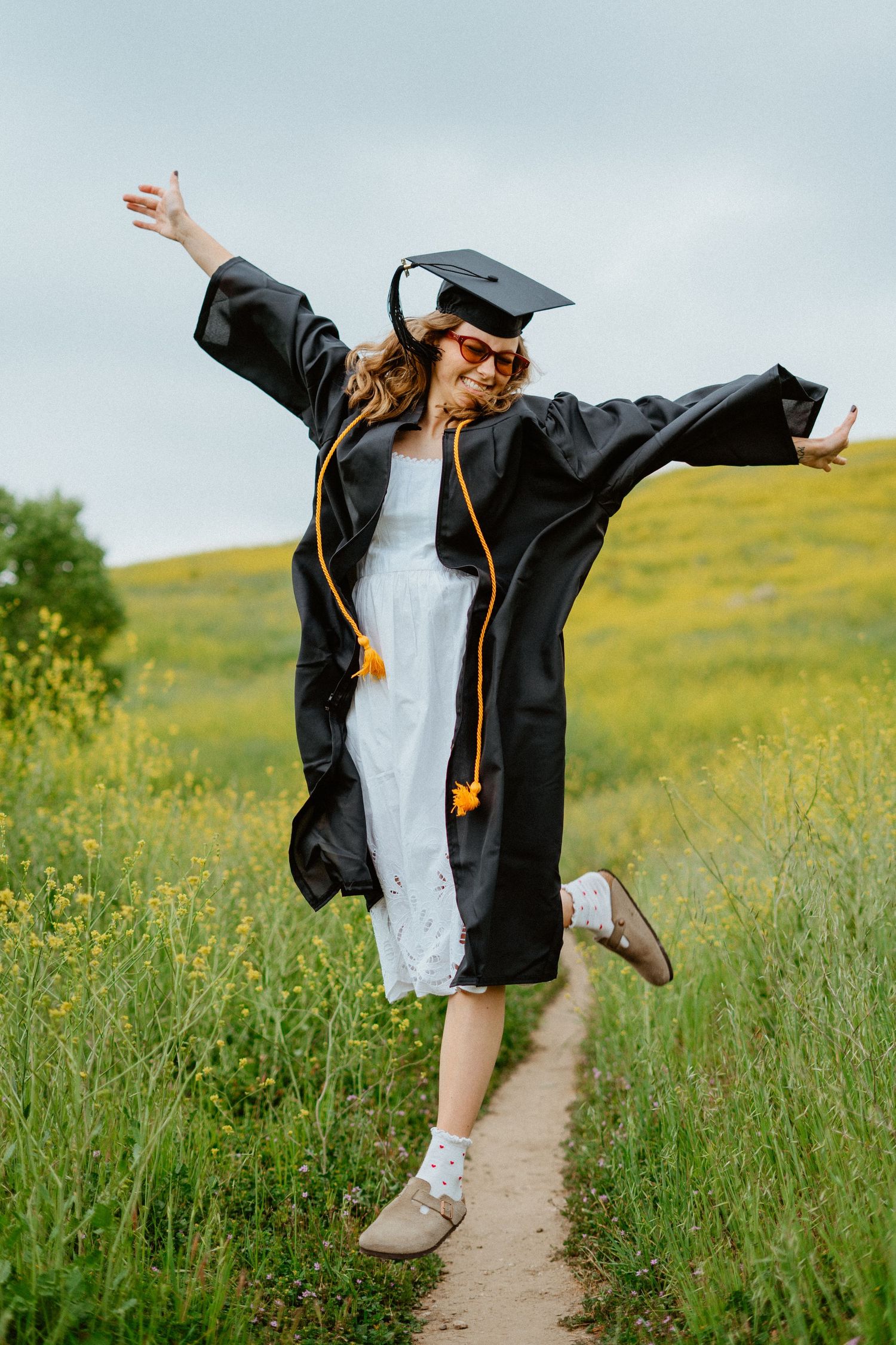 Joyful graduate in black cap and gown jumping on a wildflower meadow path.