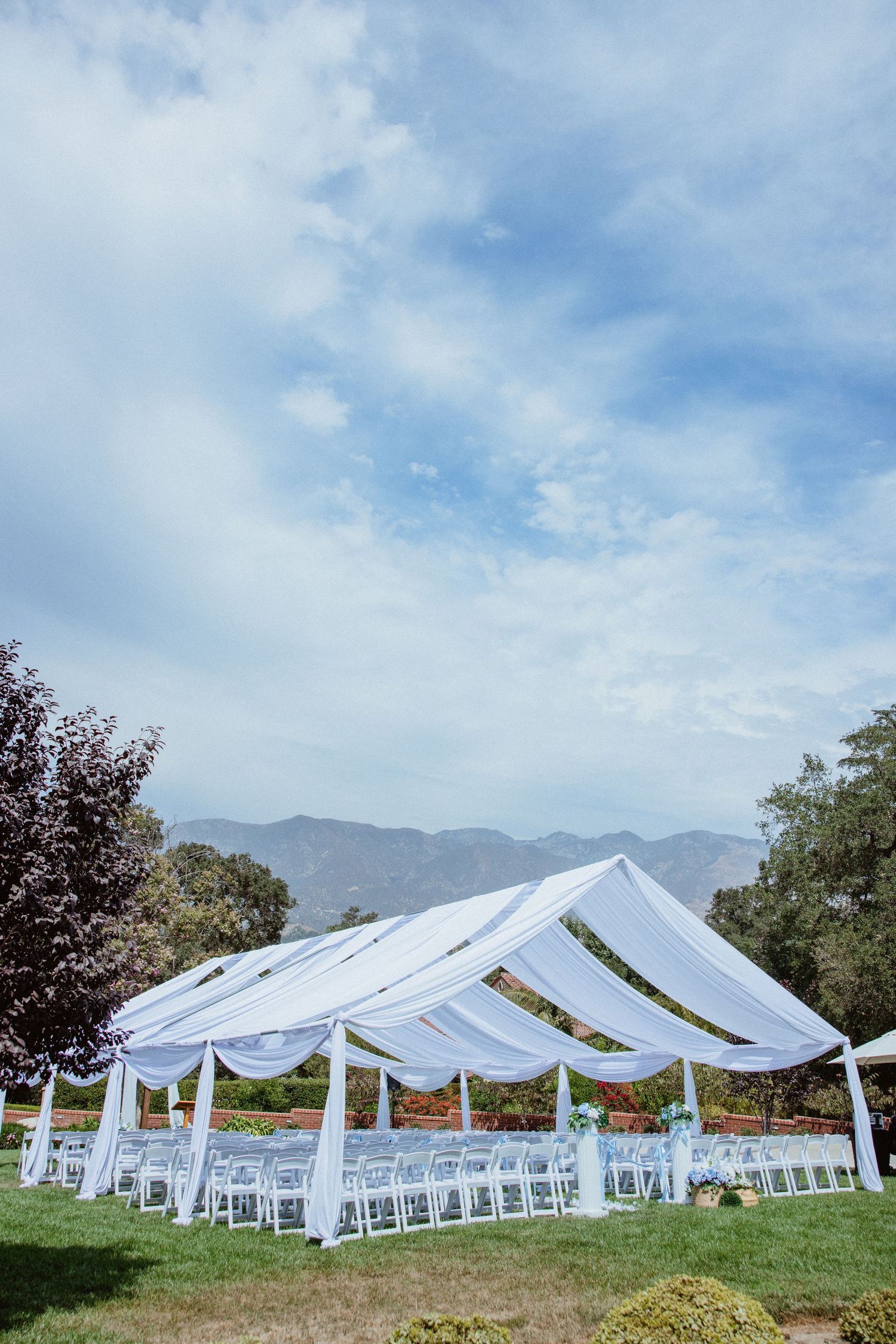 White draped wedding ceremony tent with rows of chairs set against mountain backdrop under cloudy blue sky.