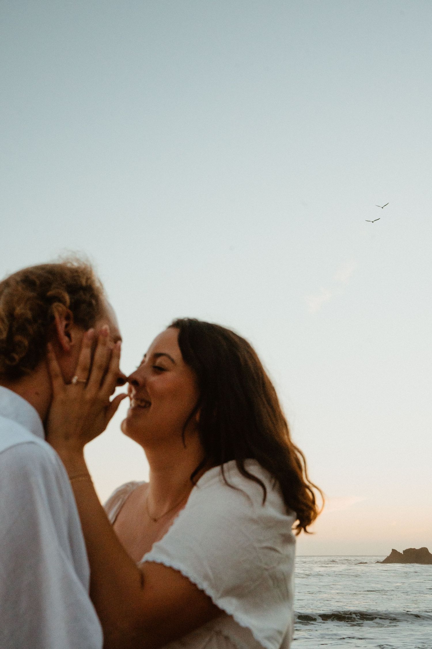A couple shares a romantic kiss at sunset on the beach, with ocean waves and a clear sky in the background.