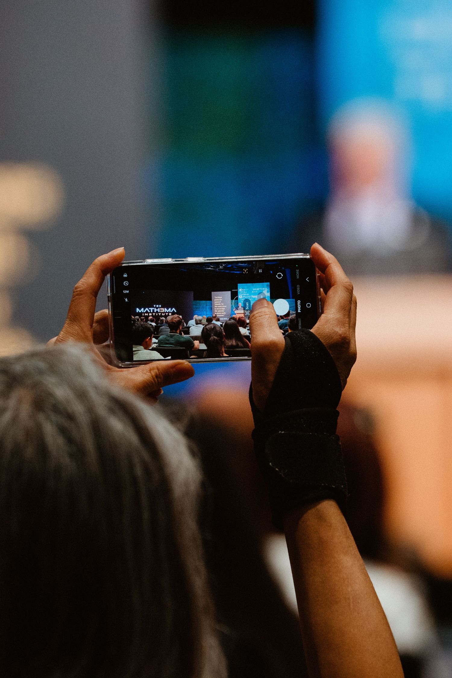 Person holding smartphone horizontally to photograph a conference presentation in a crowded auditorium.