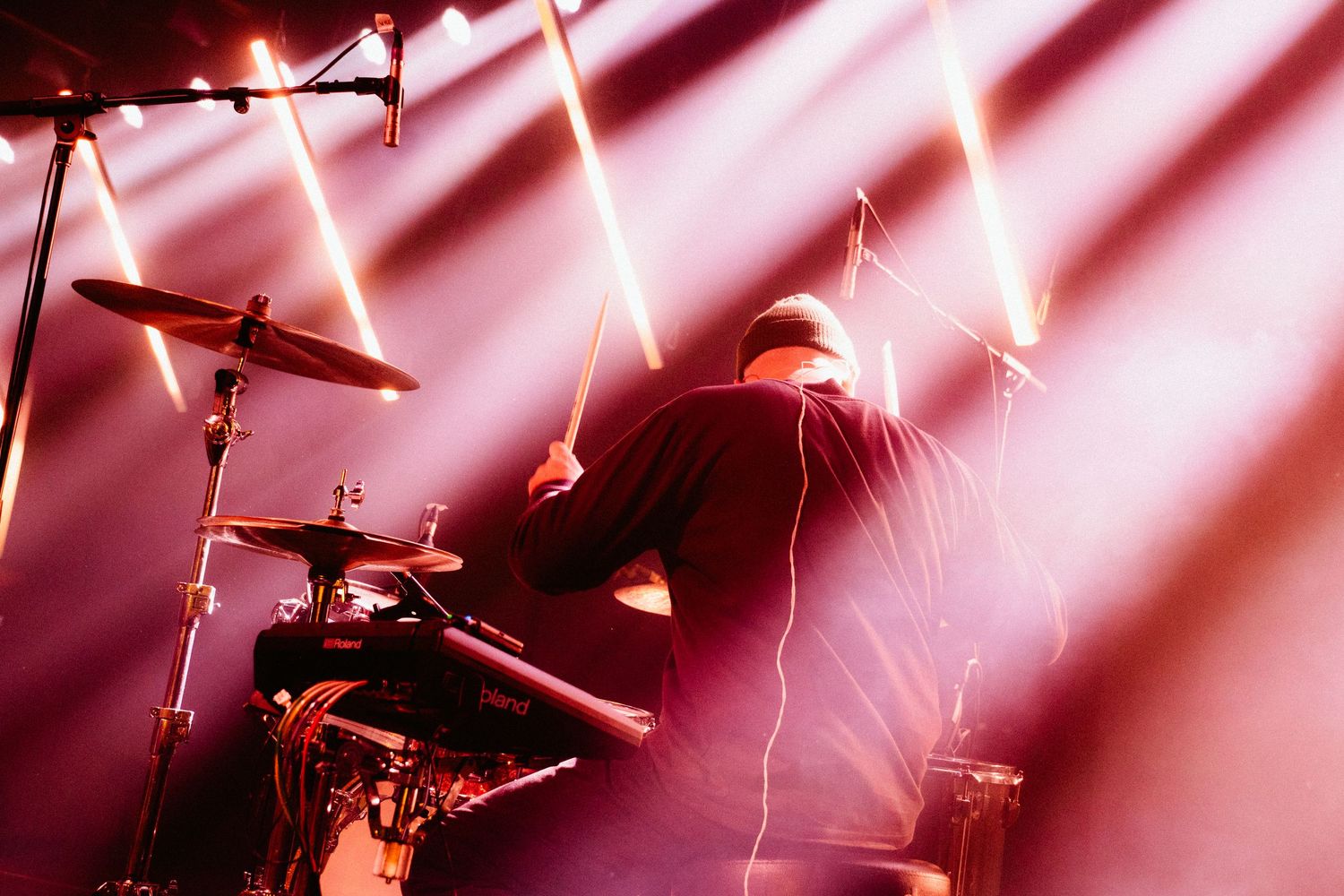 A drummer performs on stage under red and pink spotlights with dramatic rays of light.