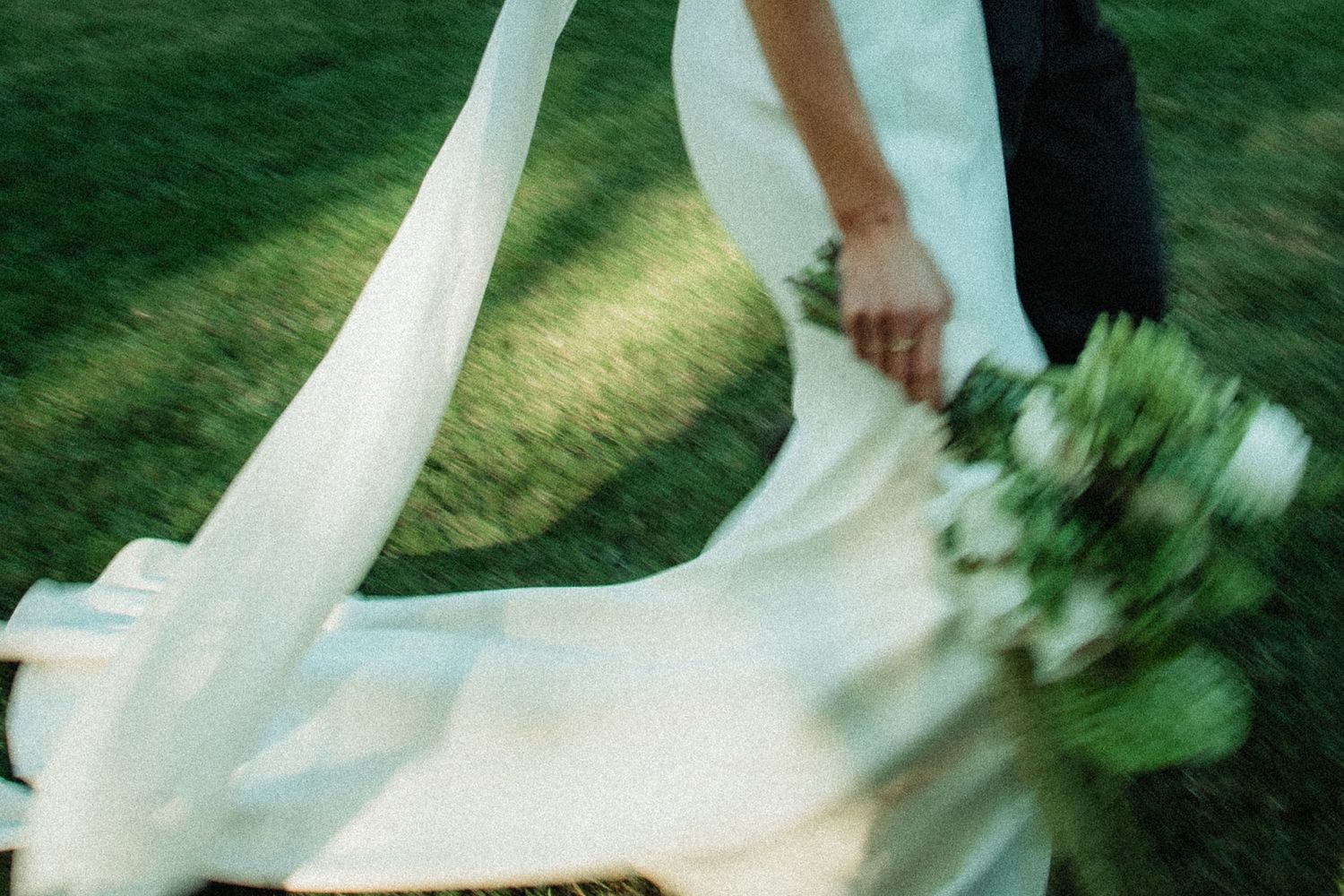 Bride in flowing white gown and veil walking with groom across green grass holding bouquet.