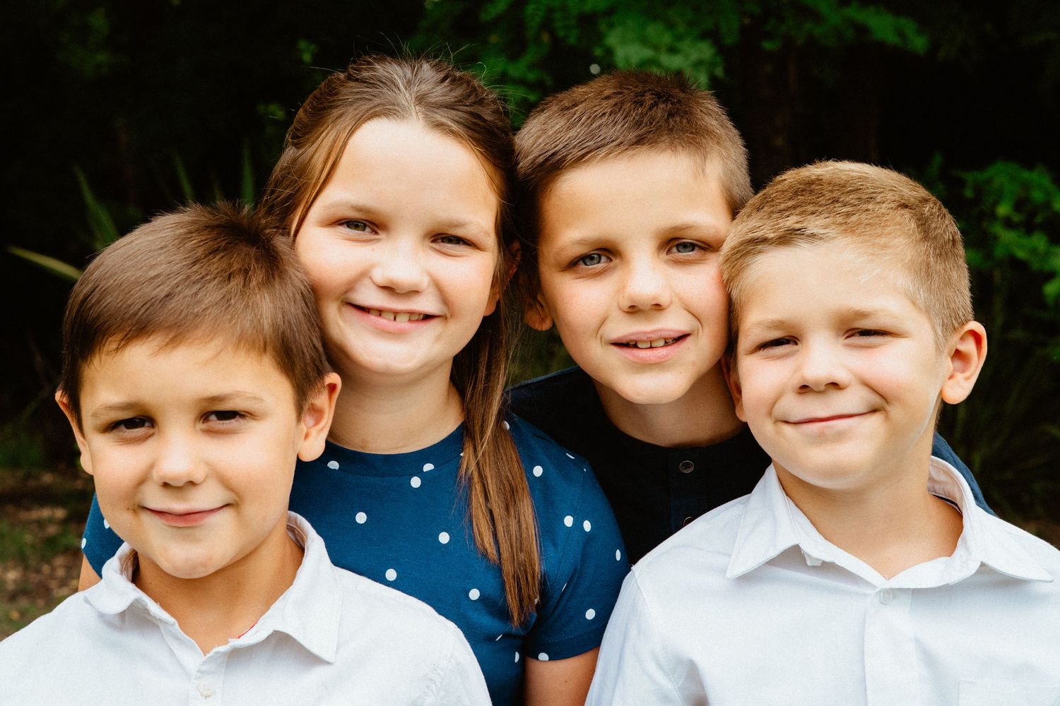 Portrait of four smiling children posing together outdoors near trees.