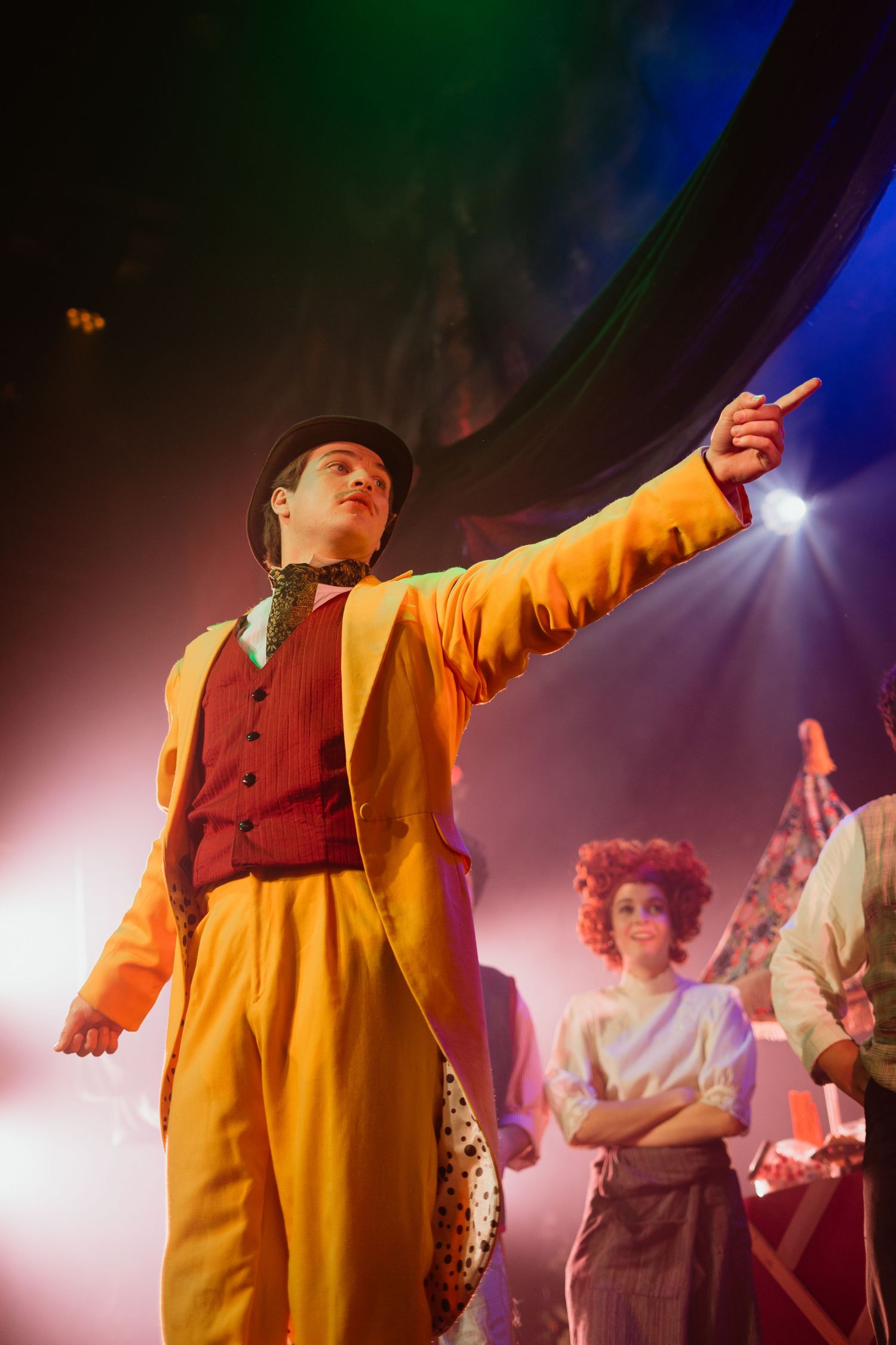 A performer in a bright yellow suit points dramatically on a colorful theatrical stage.