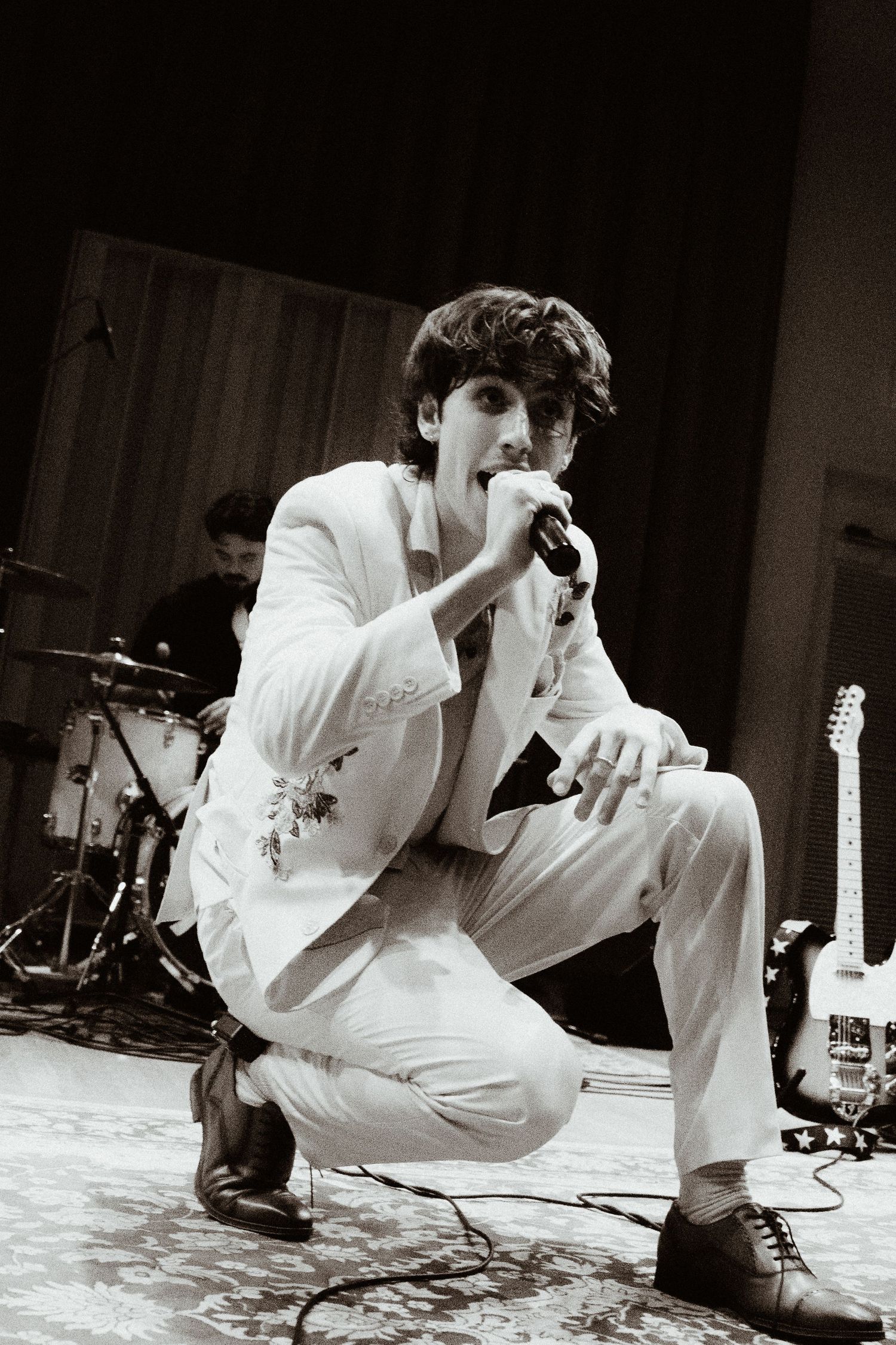 Singer in white suit crouching on stage holding microphone during live performance in black and white.