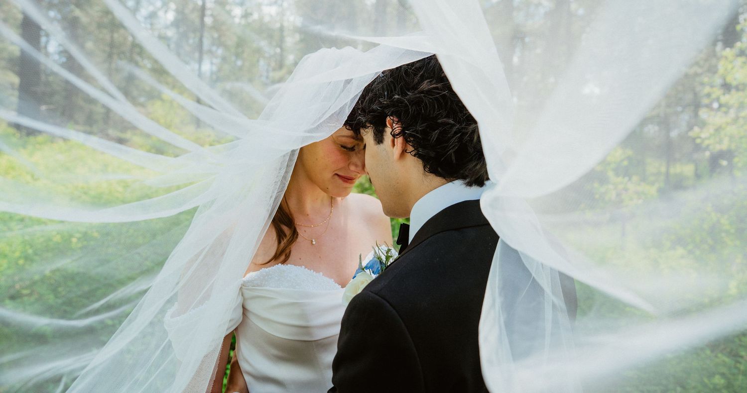 Bride and groom sharing a romantic moment under a flowing white veil outdoors on their wedding day.