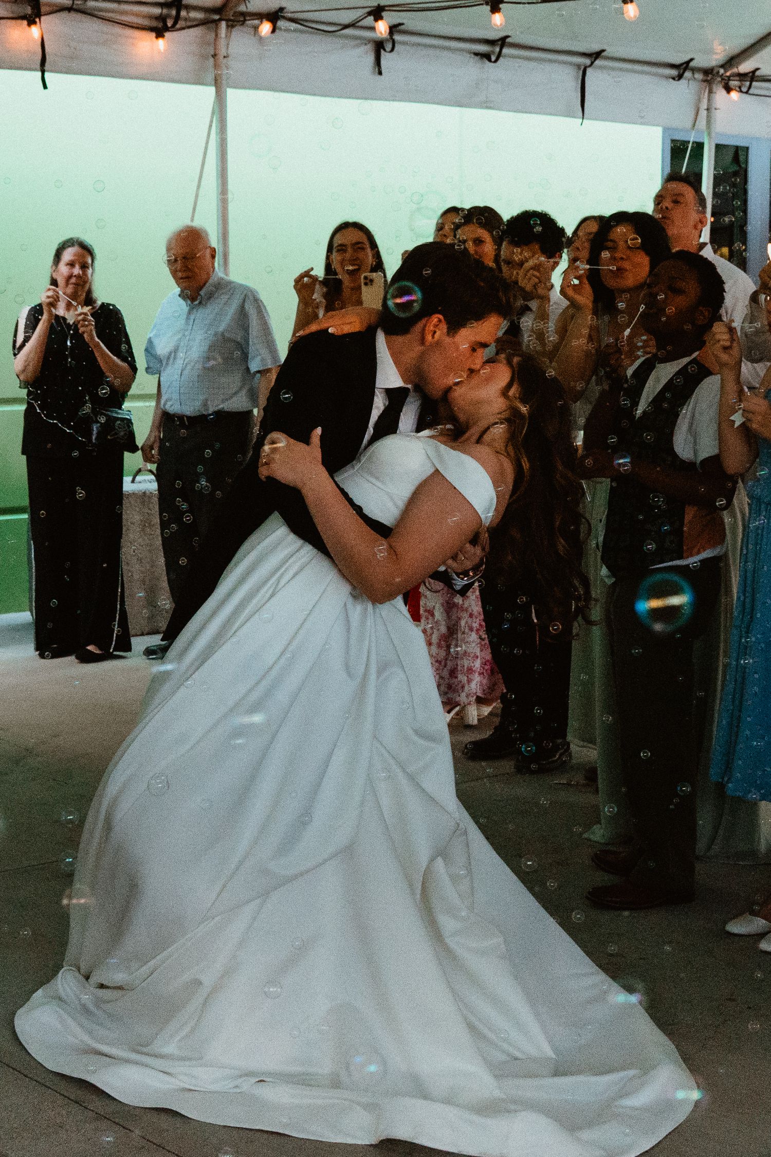Bride and groom share a romantic dip kiss on the dance floor surrounded by cheering wedding guests.