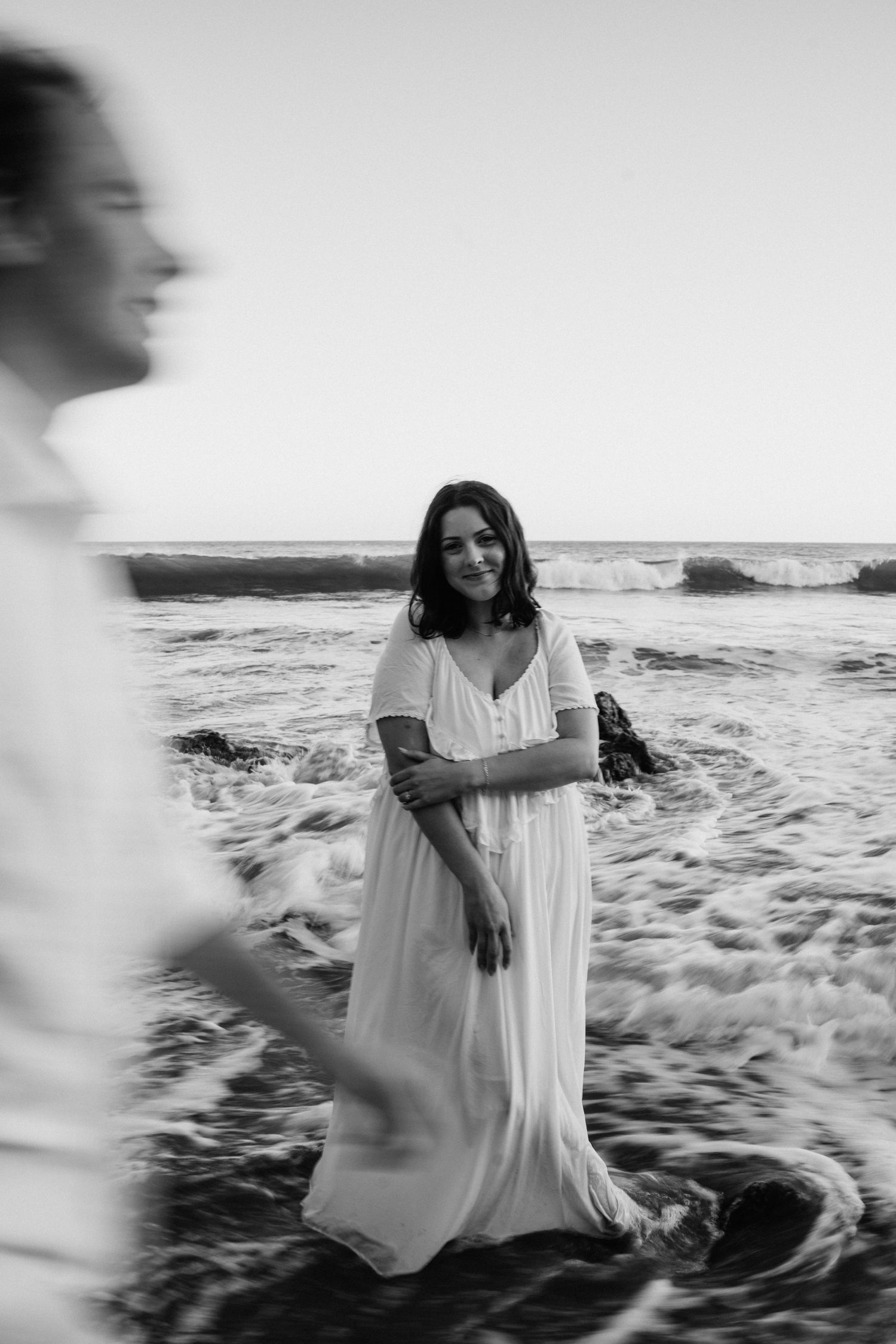 Black and white photo of a woman in a white maxi dress standing in ocean waves on a beach.
