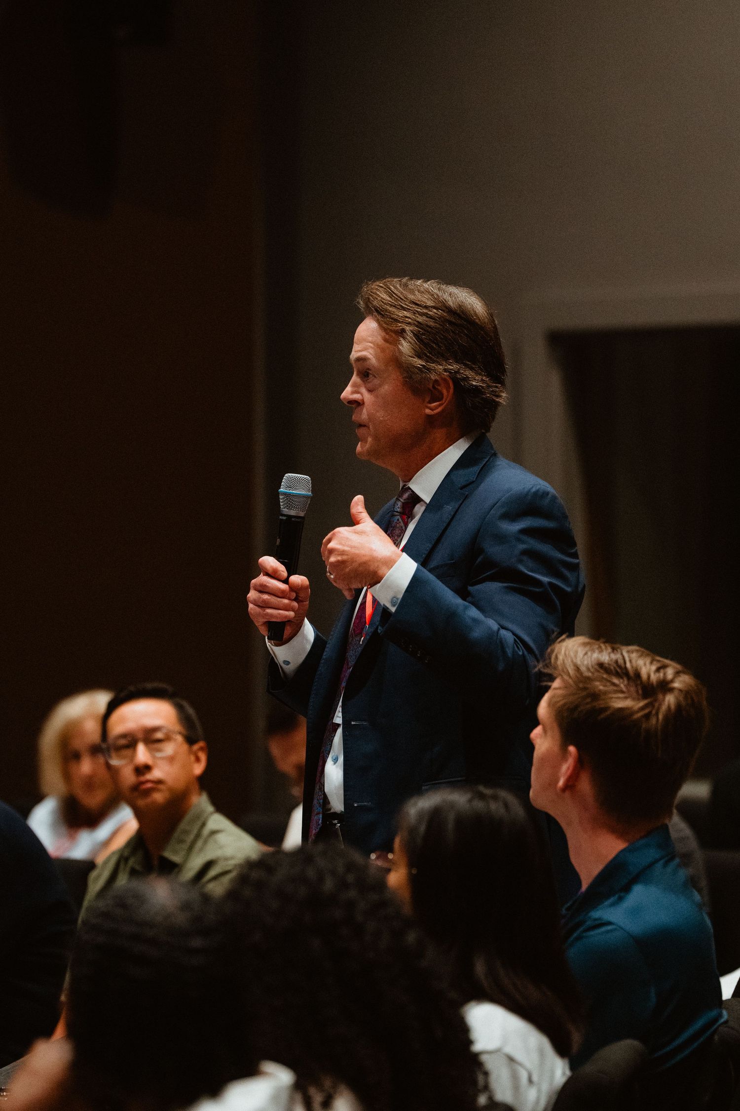 Man in blue suit speaking into microphone at a conference event with diverse audience seated in background.