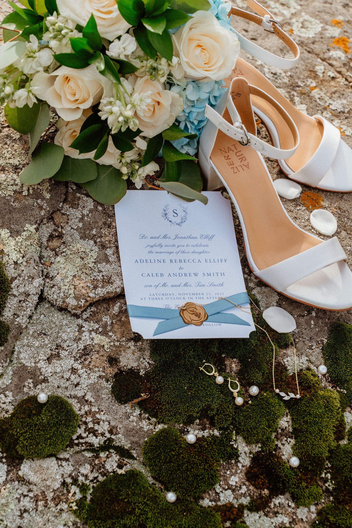 Elegant wedding invitation with blue hydrangea bouquet, white heels, and pearl jewelry on moss.