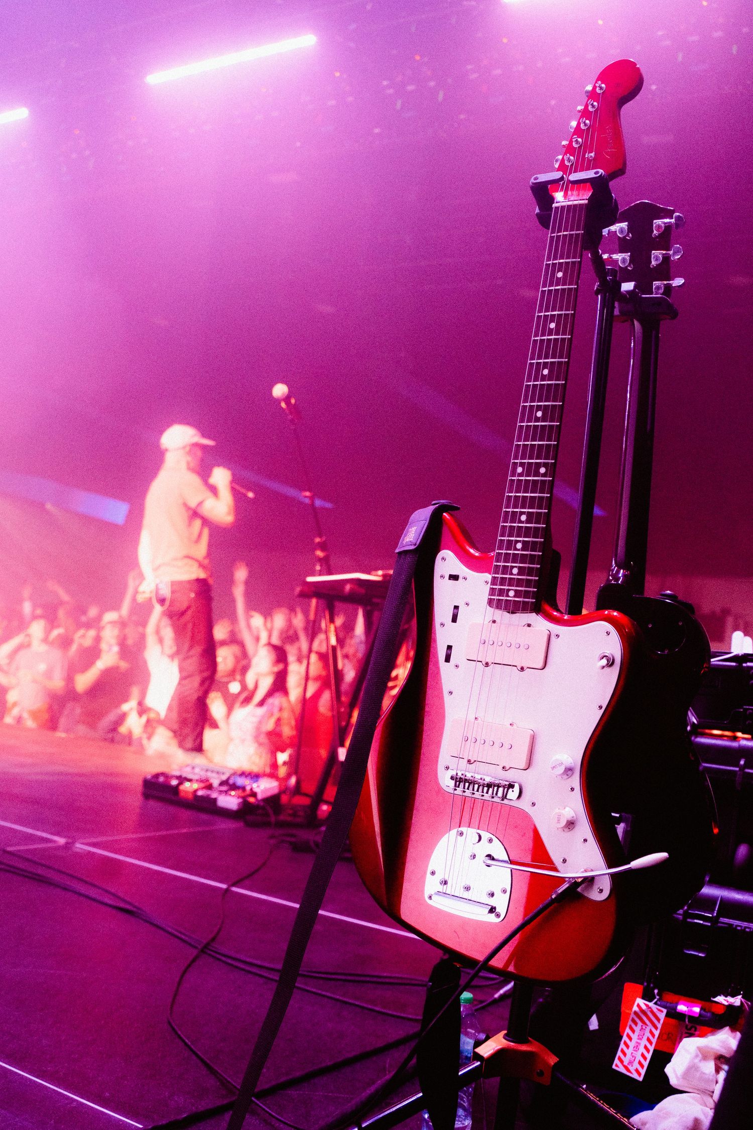 A red and white electric guitar in the foreground as a musician sings to the crowd in the background, purple lighting throughout.
