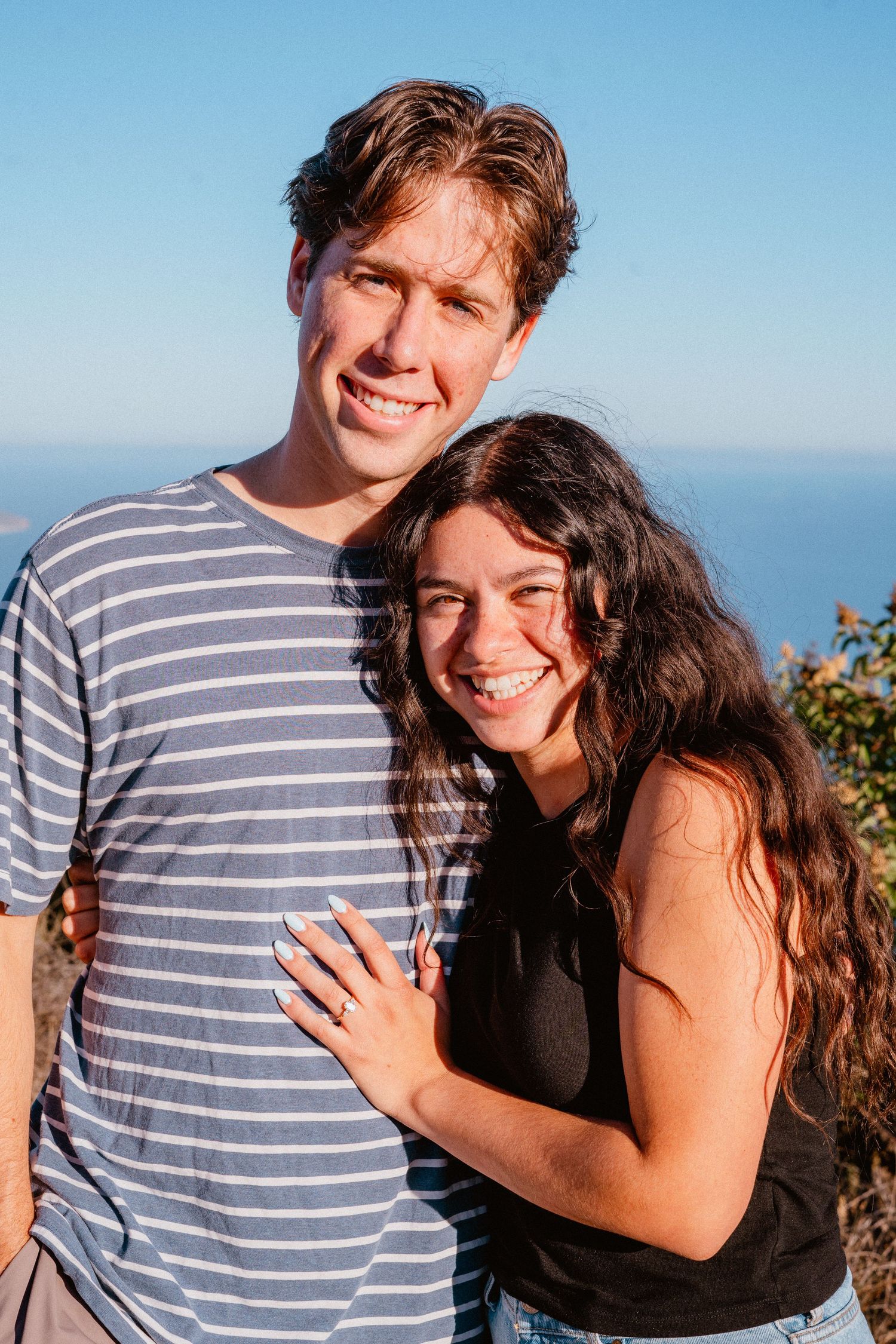 Happy smiling couple posing together outdoors with a bright blue ocean and sky in the background.