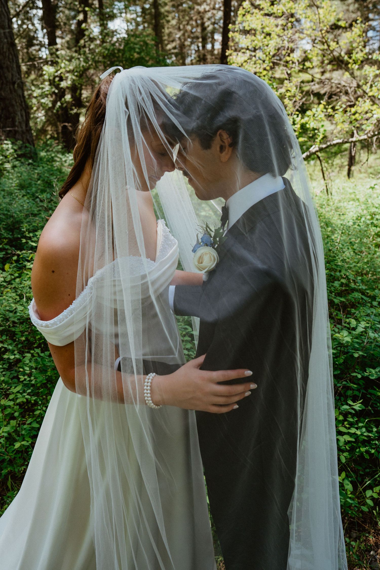 Bride and groom sharing a kiss under a flowing white wedding veil surrounded by lush green forest.