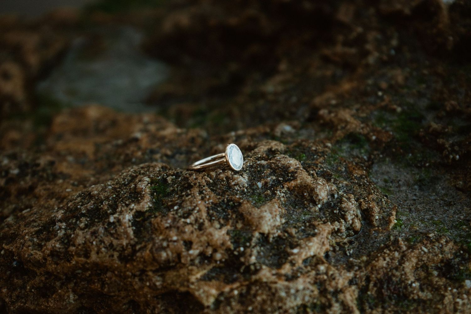 A delicate silver ring with an oval white stone resting on a textured rocky surface outdoors.