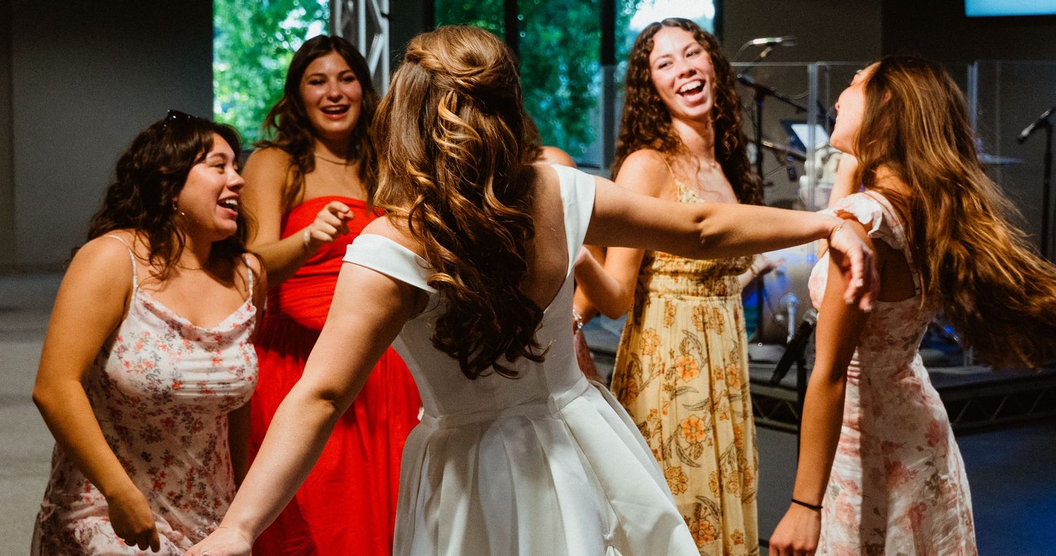 Bride in white dress laughs and dances joyfully with bridesmaids in colorful dresses at wedding reception.