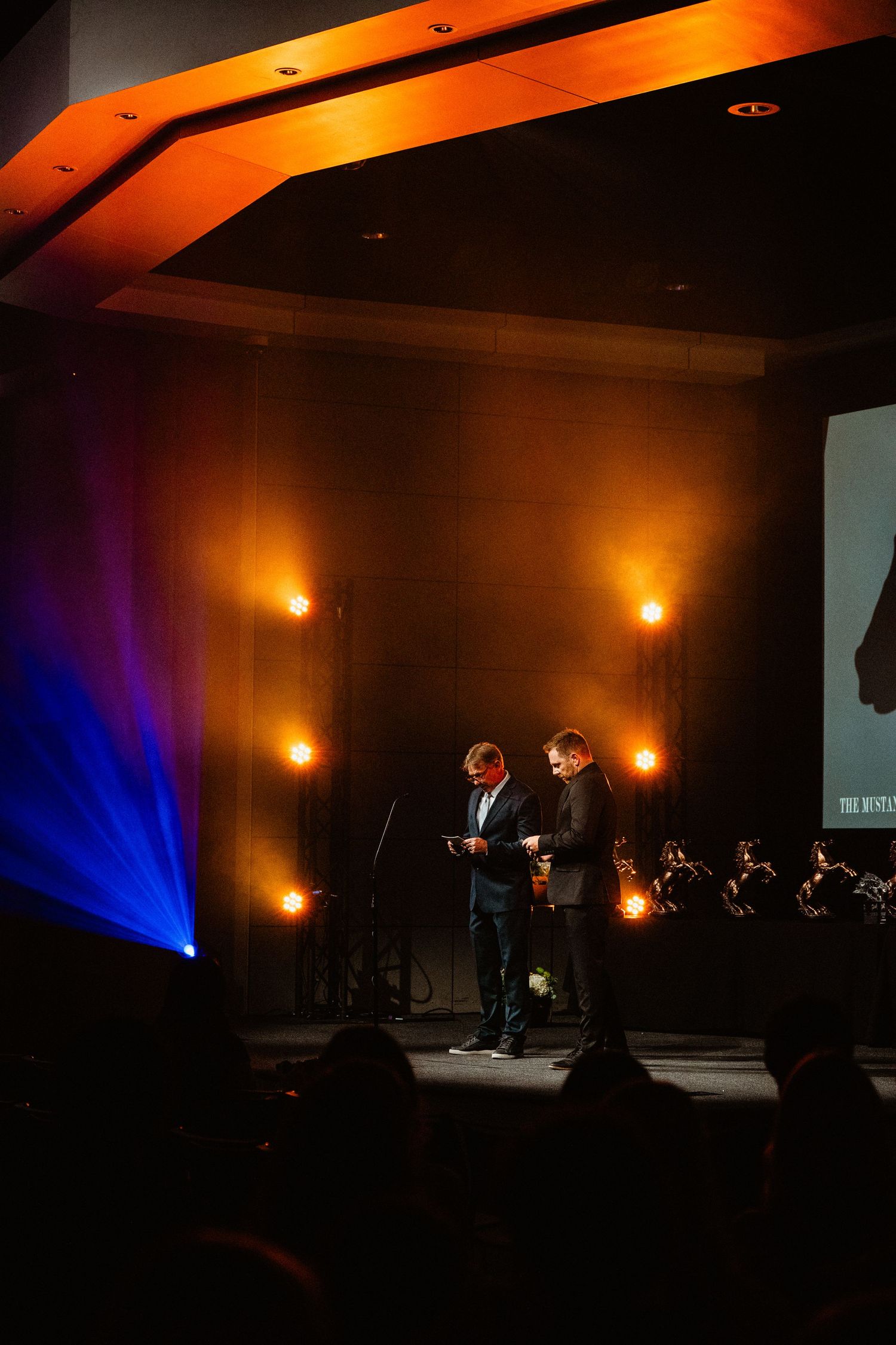 Two men present awards on a dramatic stage with warm orange uplighting and an orchestra in the background.