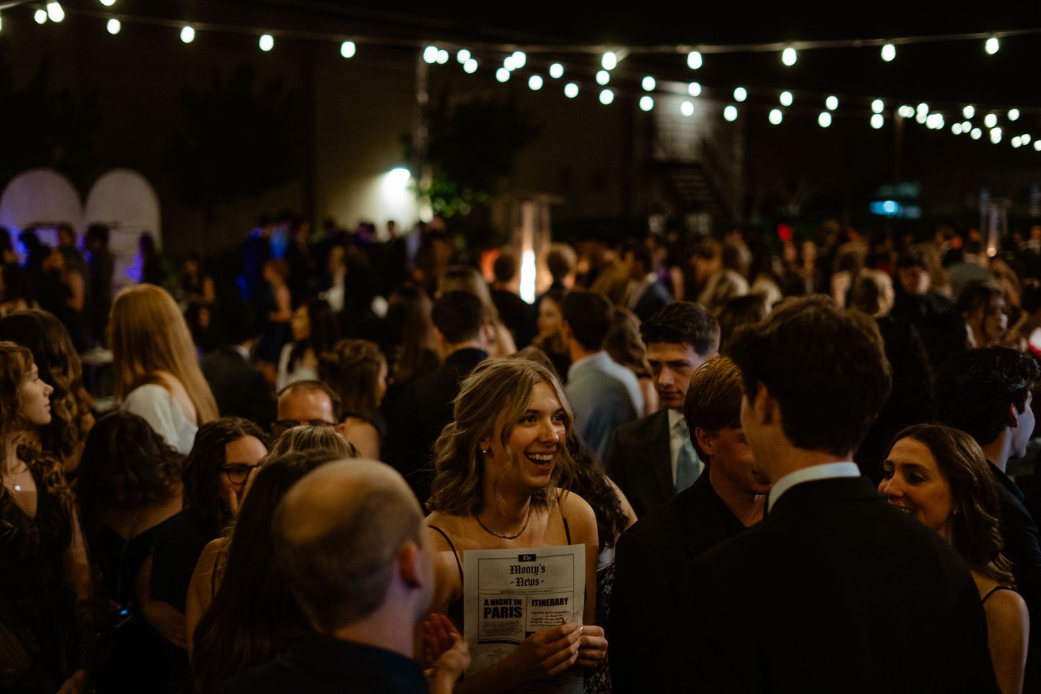 Crowd of guests socializing outdoors at evening event under string lights.