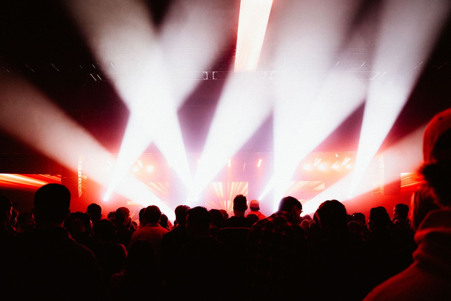 Silhouetted concert crowd facing a stage with powerful red and white spotlight beams.