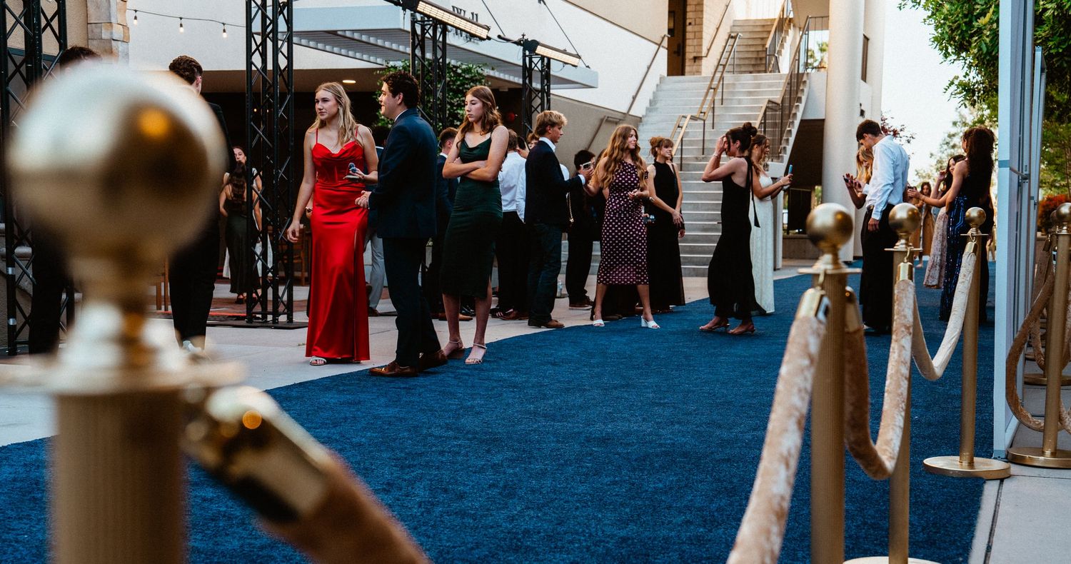 Elegantly dressed guests walk a blue carpet at a formal evening gala event with velvet rope barriers.