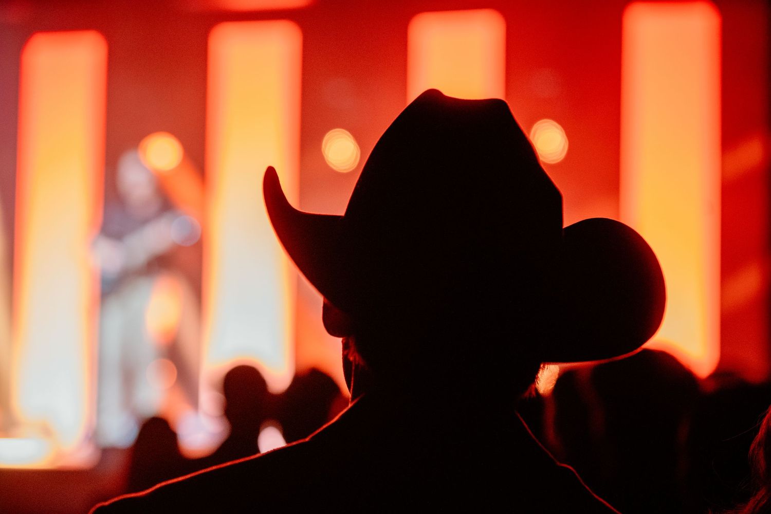 A silhouetted cowboy hat wearer watches a country music performer on a warmly lit red stage.
