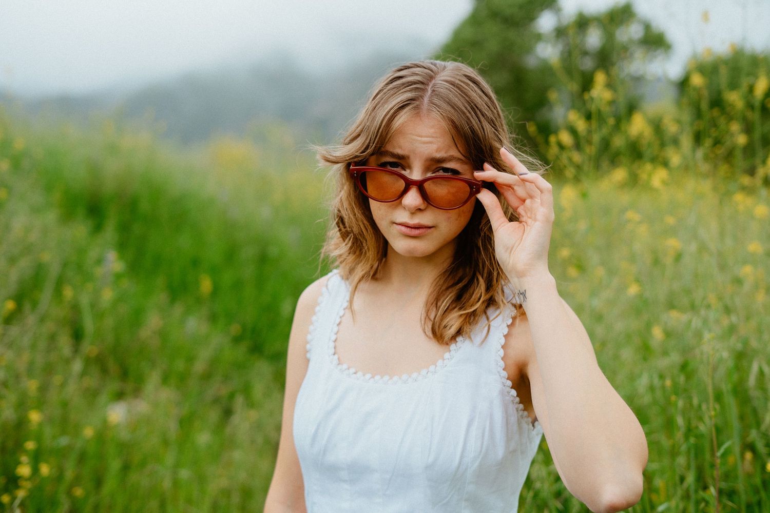 Young woman in white dress holds orange sunglasses while standing in a lush green outdoor field.