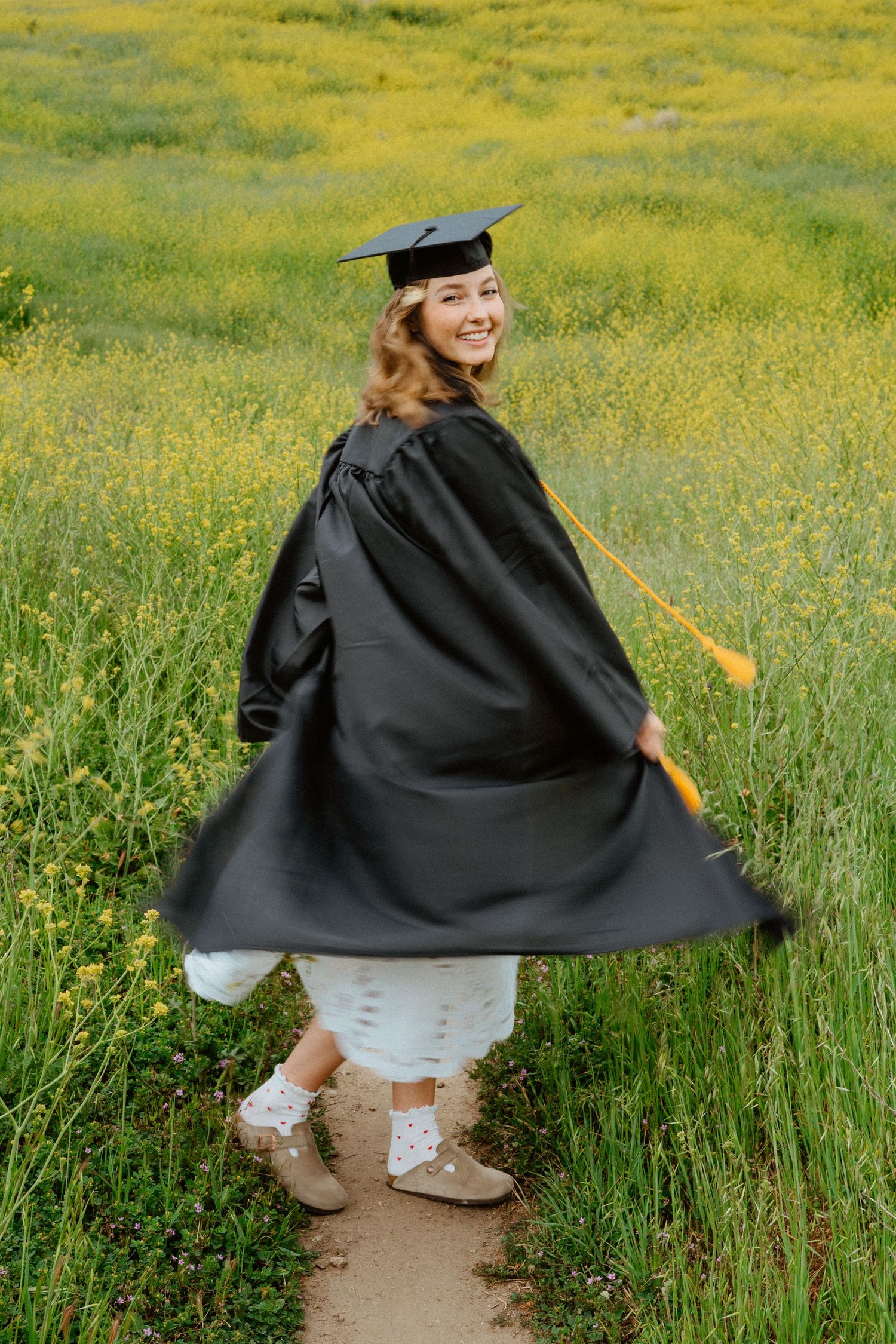 Happy graduate twirling in black cap and gown on a scenic wildflower field path.