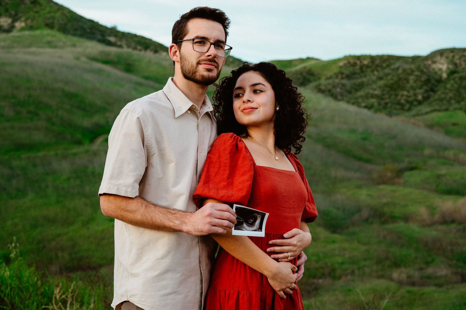 Couple standing together on green hillside holding ultrasound photo, woman in red dress smiling softly.