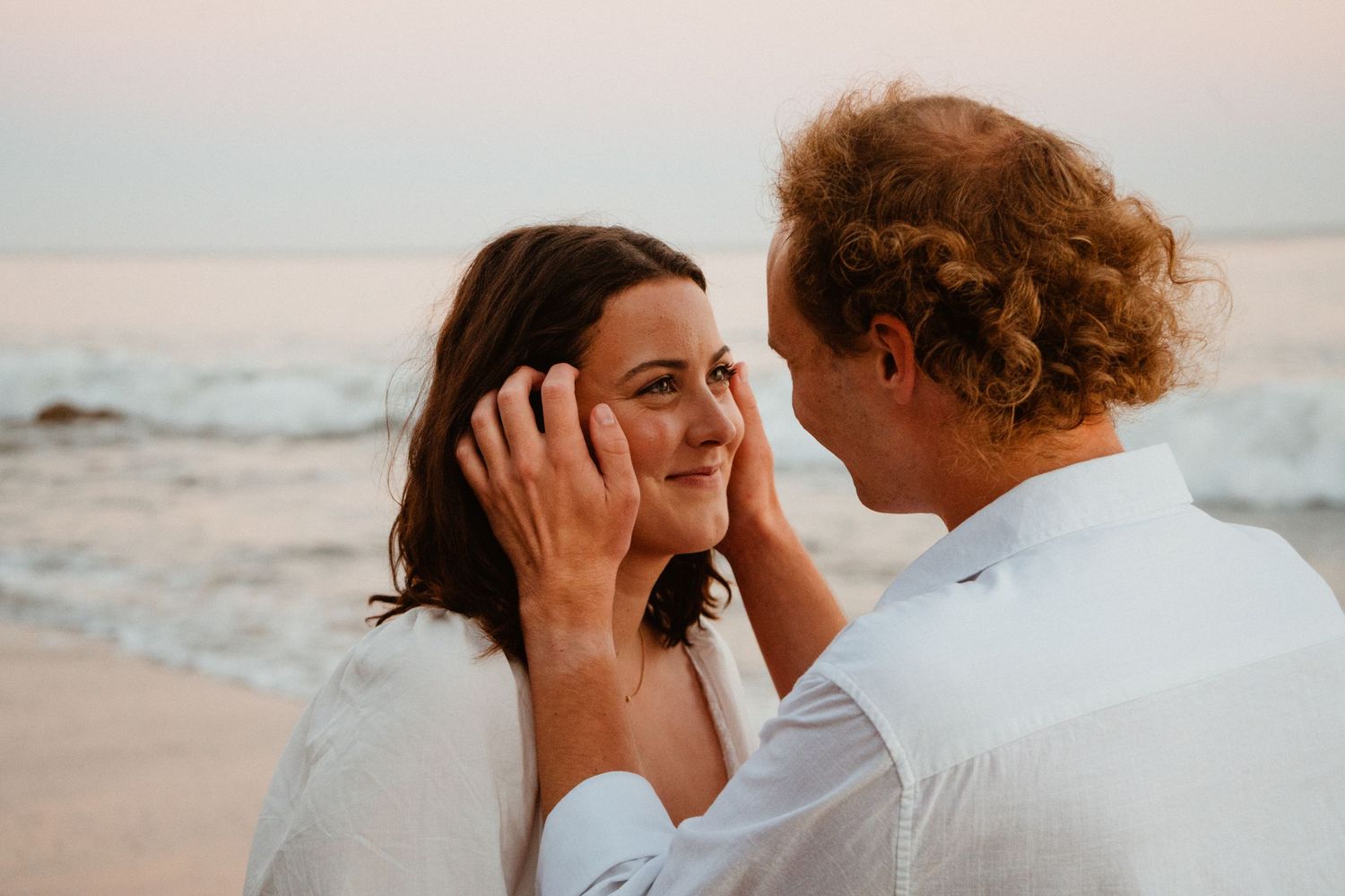 A couple shares a tender moment on the beach at sunset, with the man gently cradling the woman's face as she smiles.
