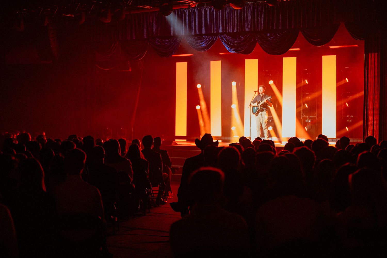 A performer on a lit stage with glowing columns entertains a silhouetted crowd at a live concert.