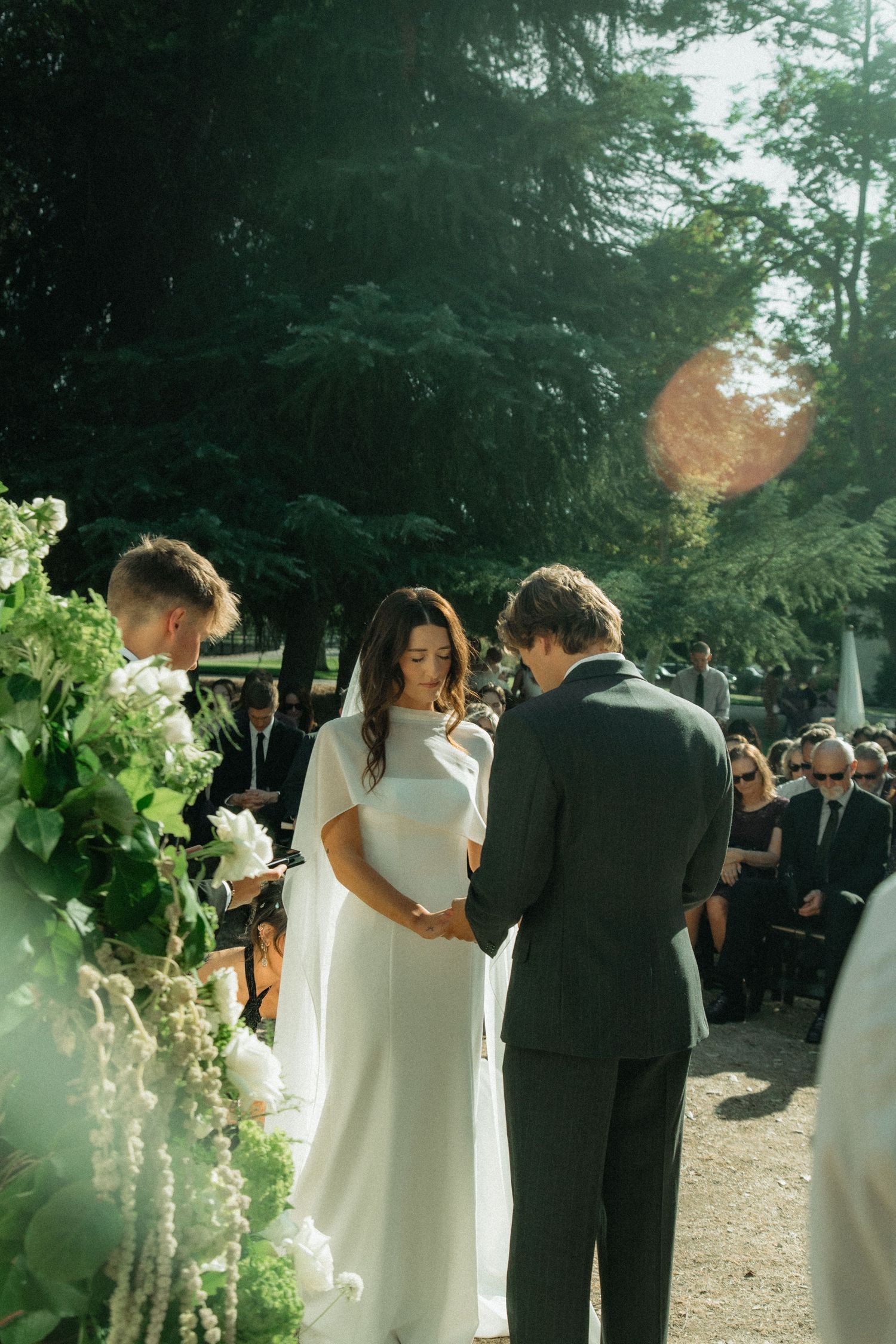 Bride and groom holding hands during outdoor wedding ceremony with guests seated among tall pine trees.