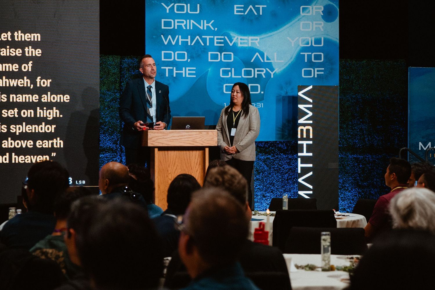 Two speakers present at a Christian conference podium with biblical scripture displayed on a blue backdrop.