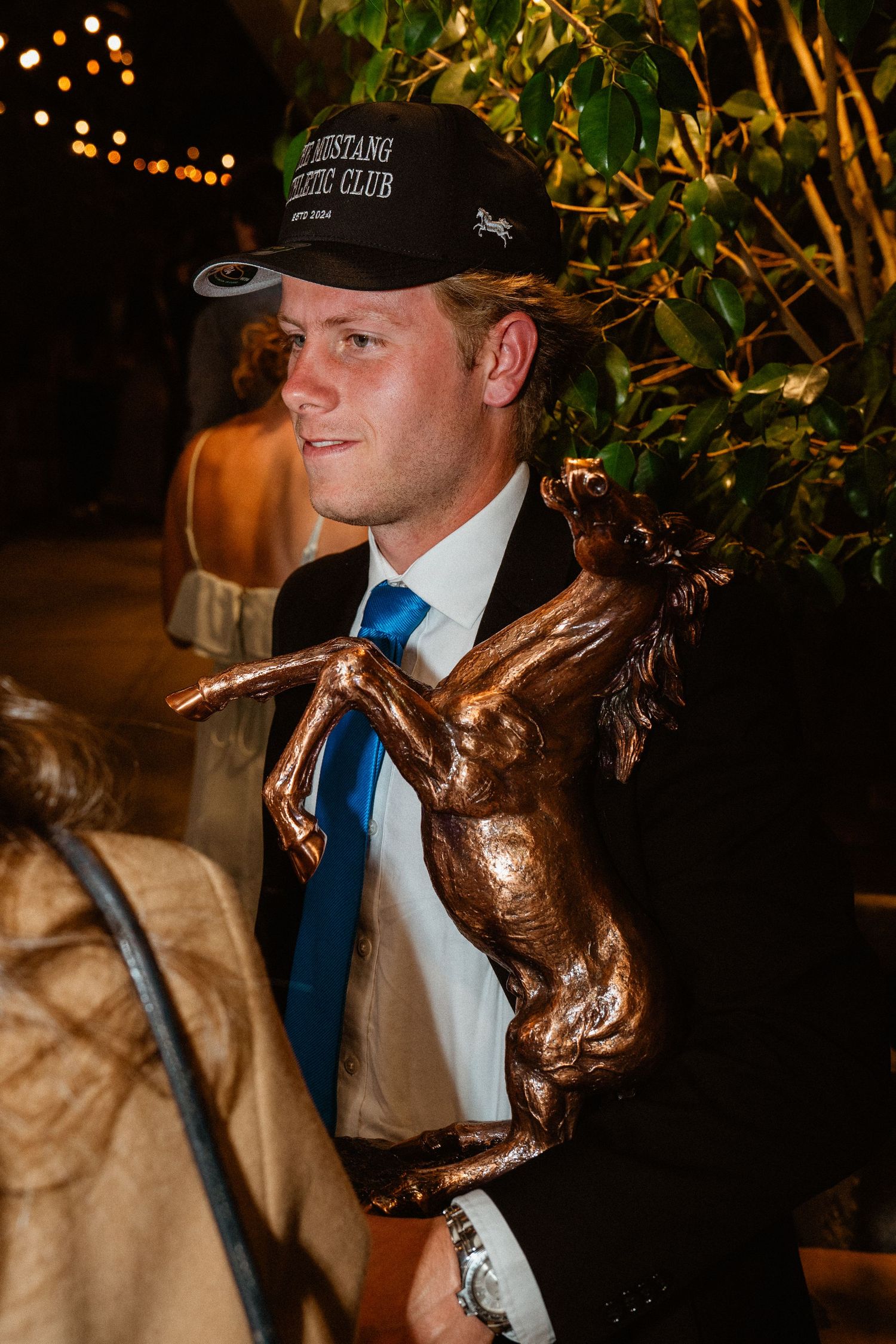 Young man in a suit and Mustang Athletic Club cap holding a bronze horse trophy at a gala.