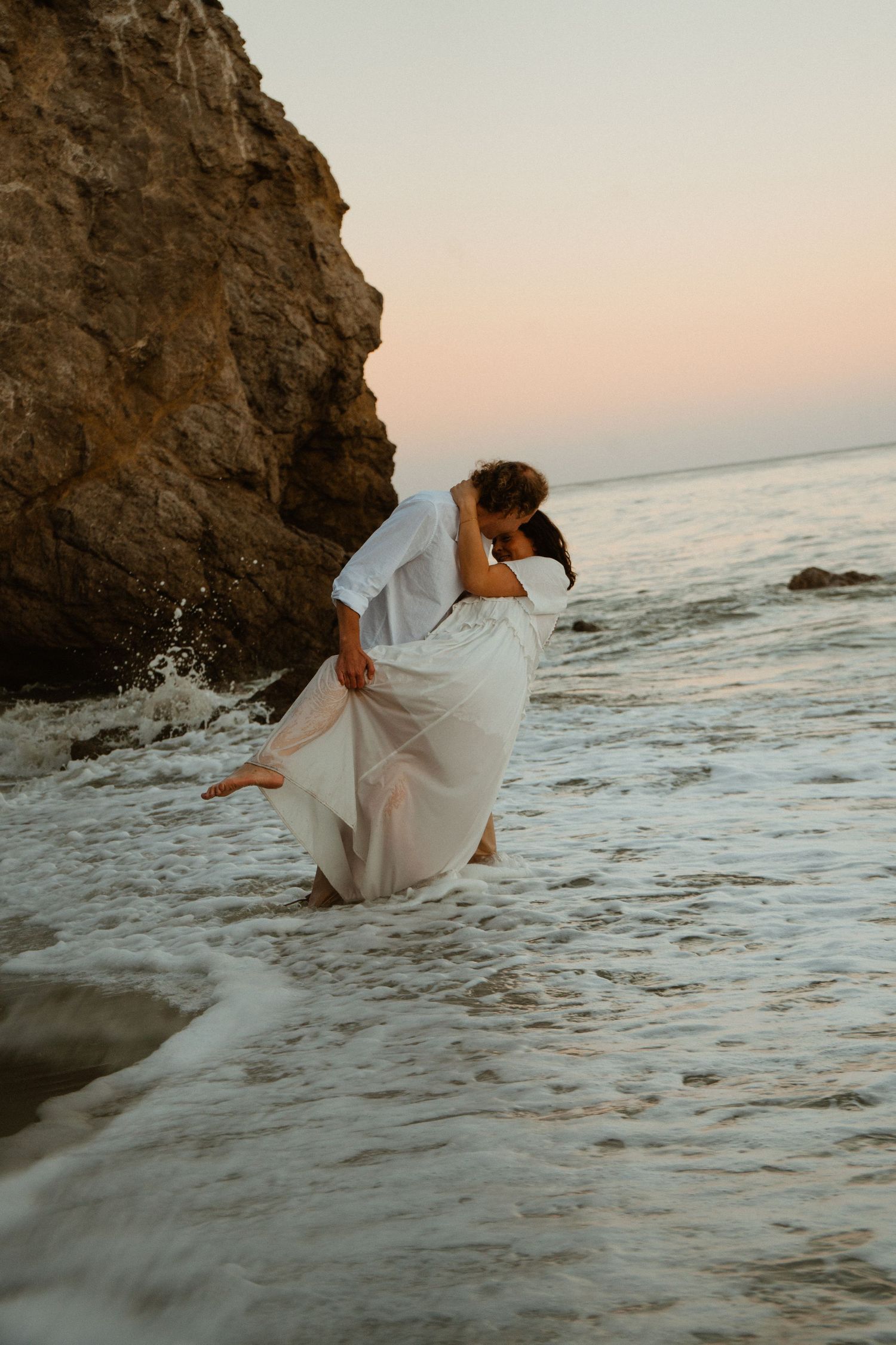 Couple sharing a romantic kiss in ocean waves at sunset, woman in flowing white dress near rocky cliffs.