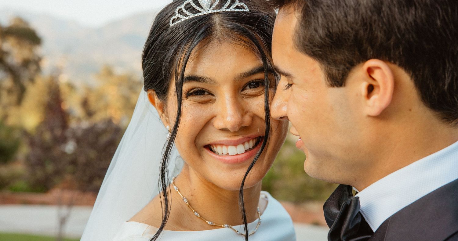 Smiling bride wearing tiara and veil gazes at groom during outdoor wedding ceremony with mountain views.
